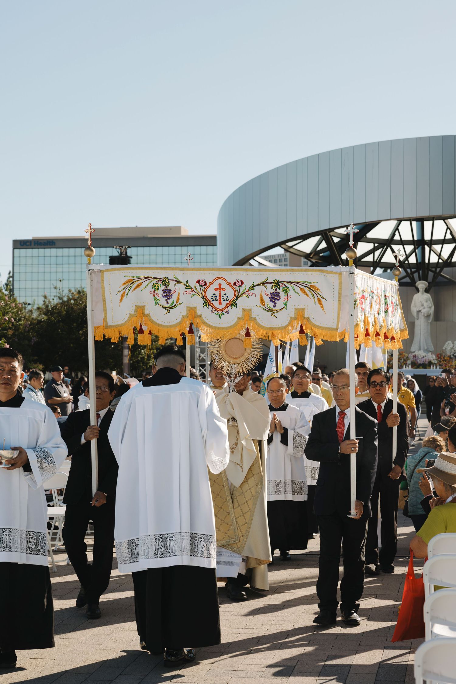 Procession of clergy and faithful during outdoor Catholic ceremony under yellow and white decorative banners.