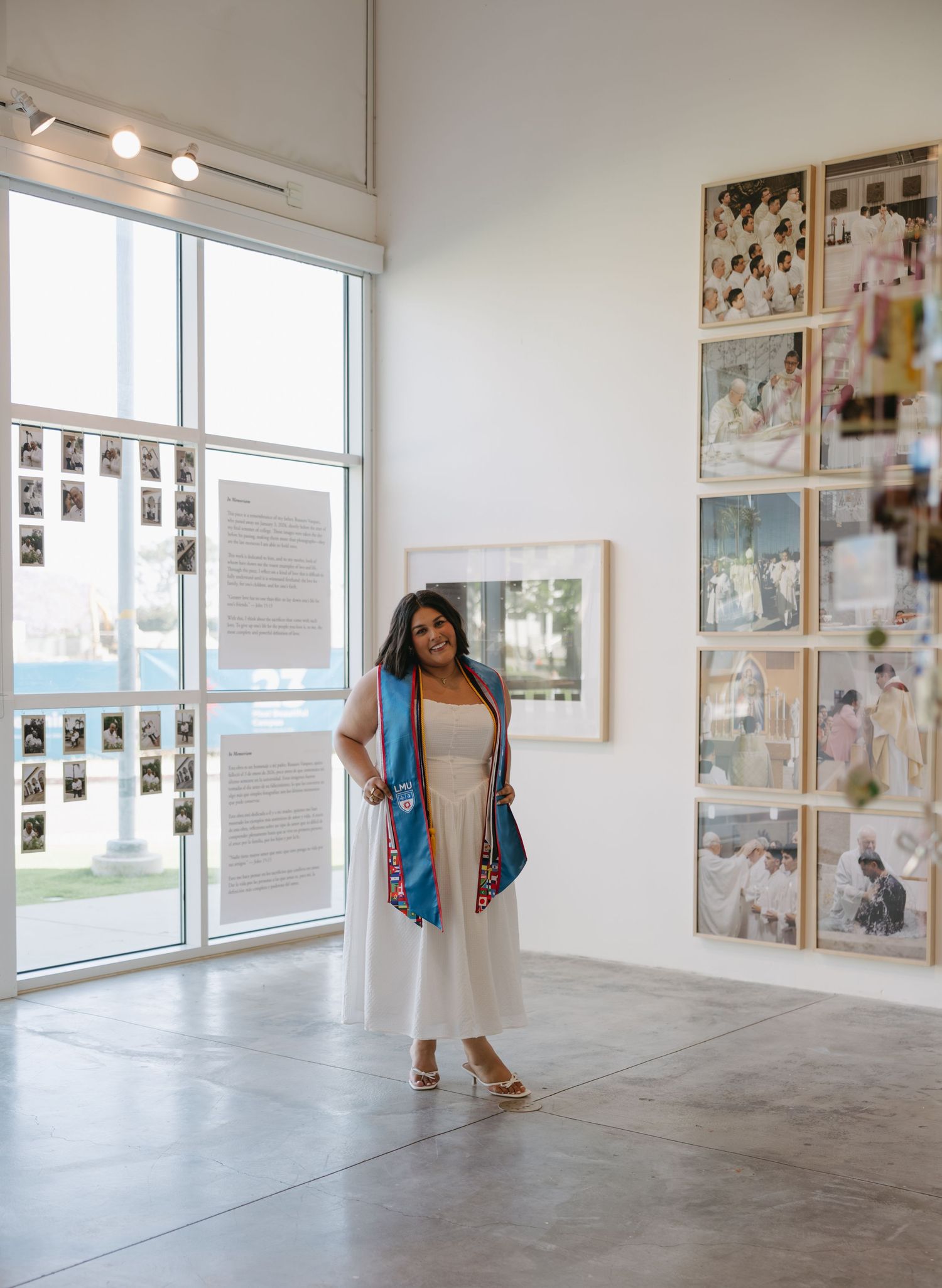 Woman in white dress and blue sash stands in modern art gallery with large windows and framed artwork.
