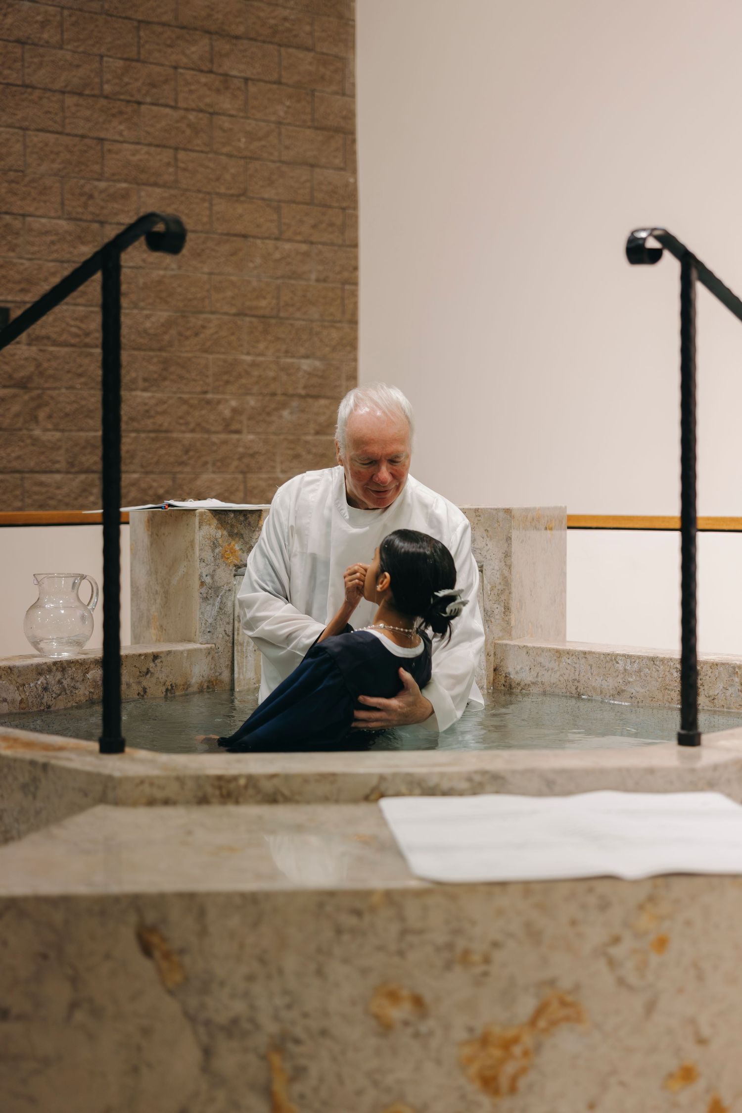 Elderly priest in white robes performing full immersion baptism of young person in baptismal pool.