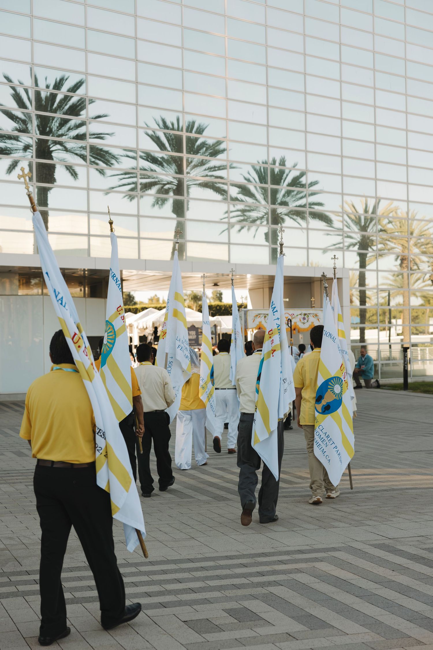 Group carrying religious banners walks past modern glass building with palm trees.