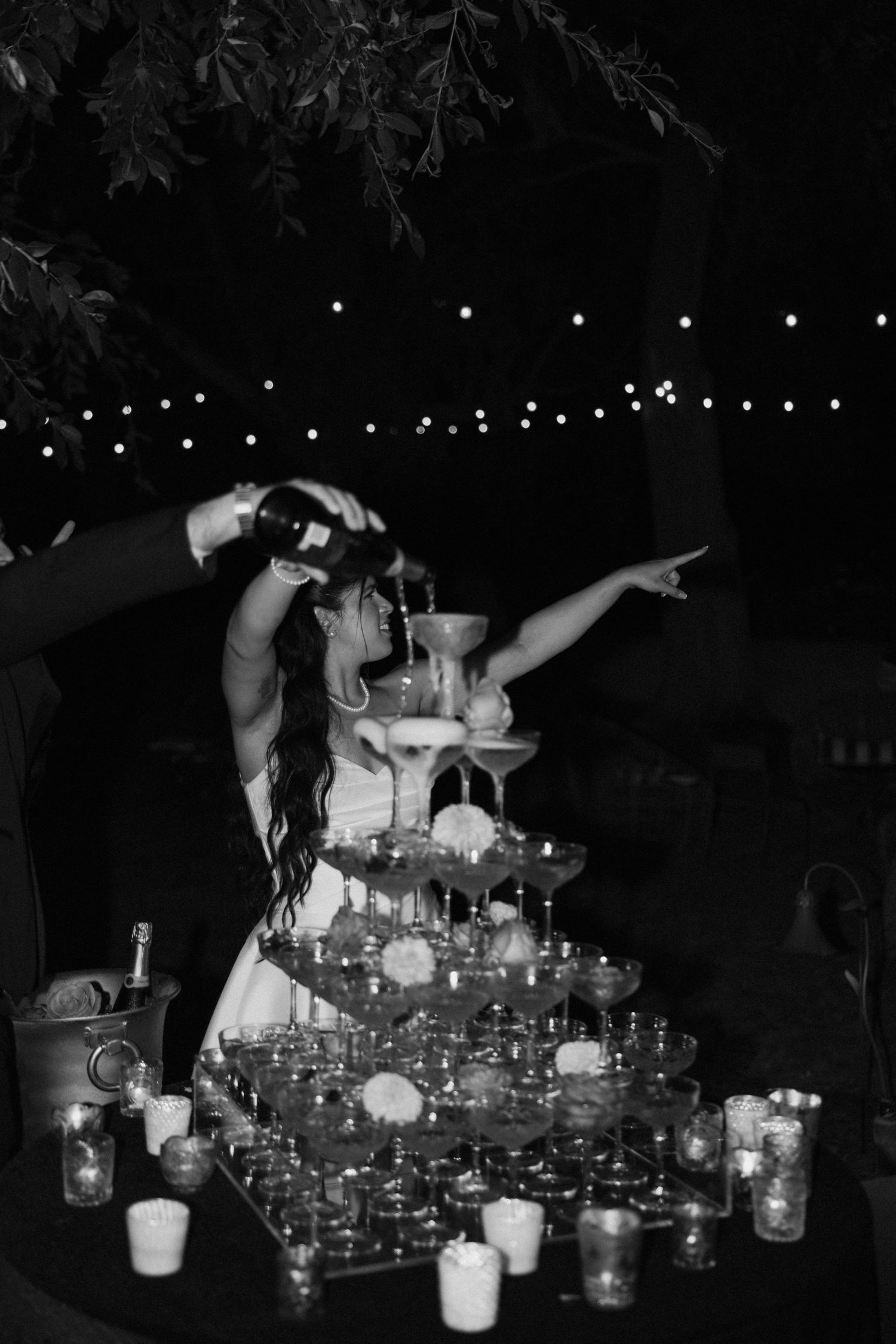 A black and white photo of champagne being poured into a tower of glasses under string lights.