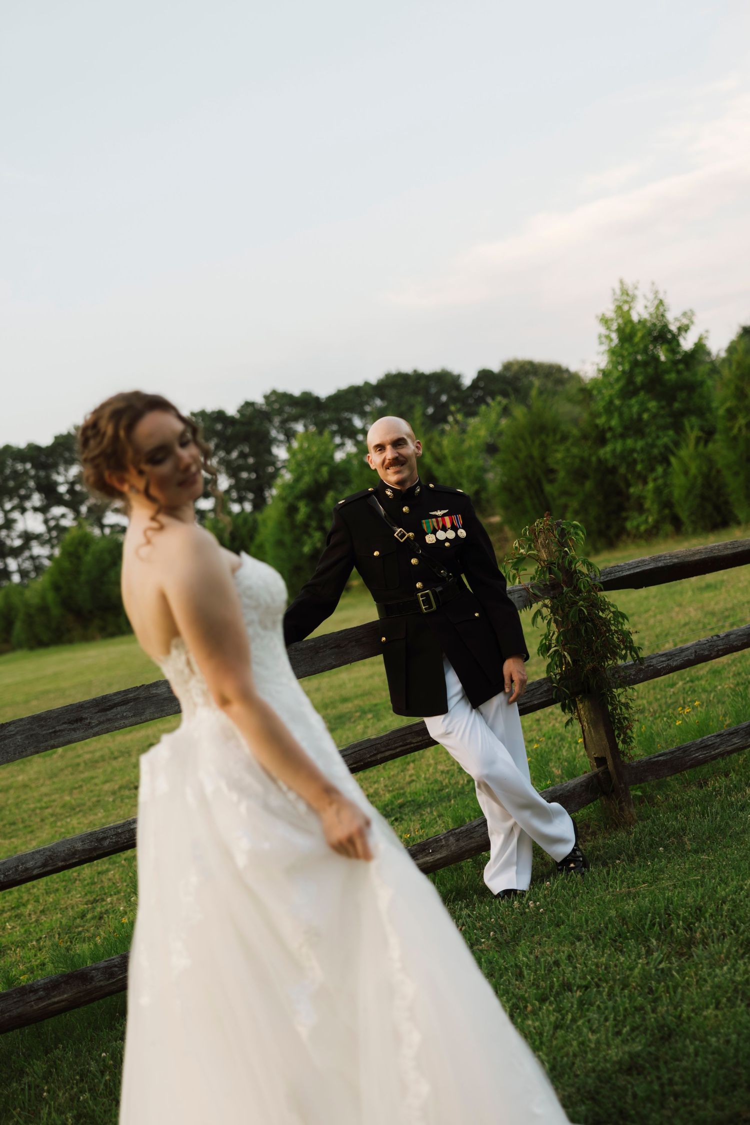 Marine groom in dress uniform smiles at bride in white wedding gown outdoors near rustic fence at sunset.