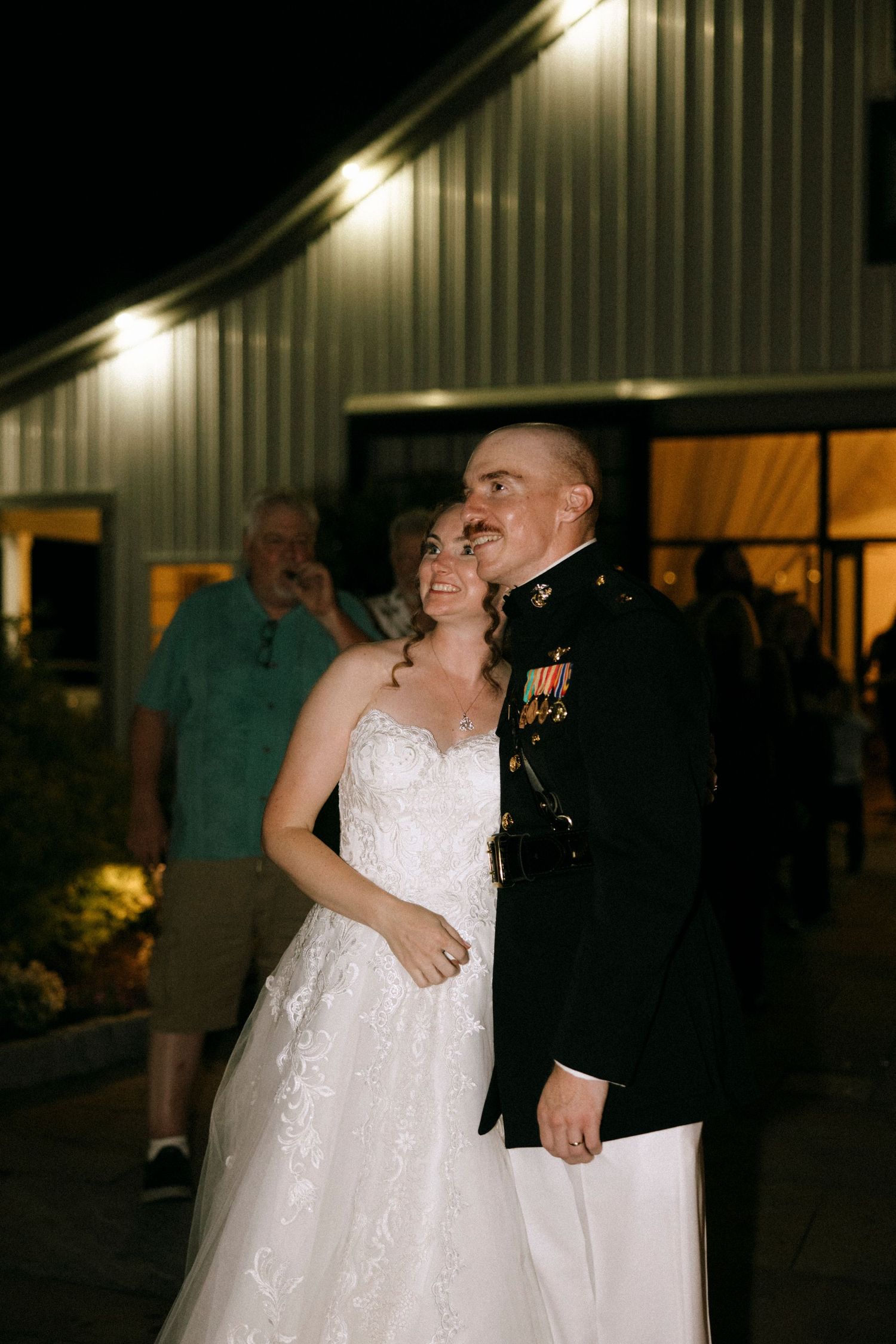 A bride in a white lace gown and groom in military dress uniform smile joyfully outside a barn venue at night.