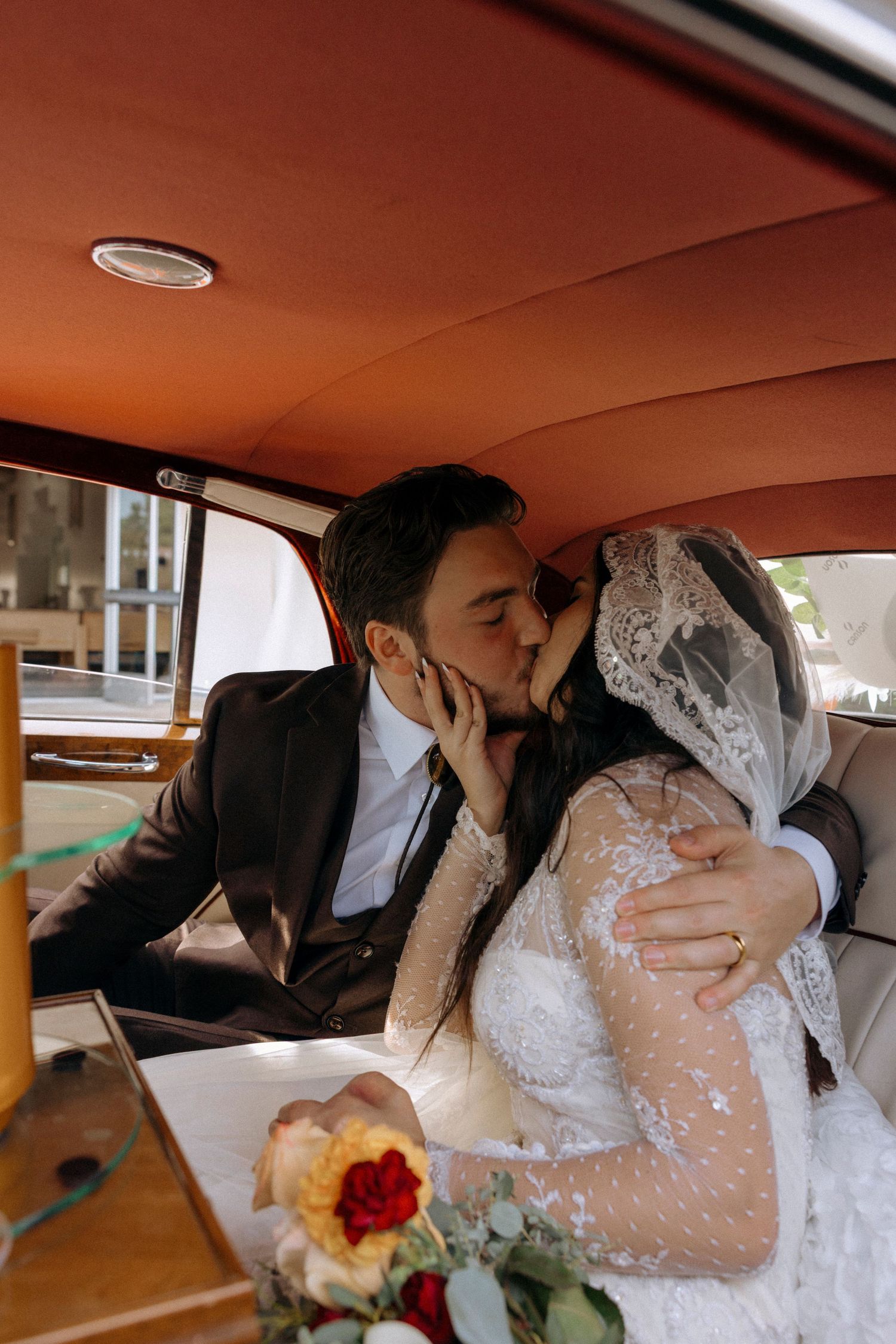 A tender wedding moment captured through the window of a classic car with warm lighting.