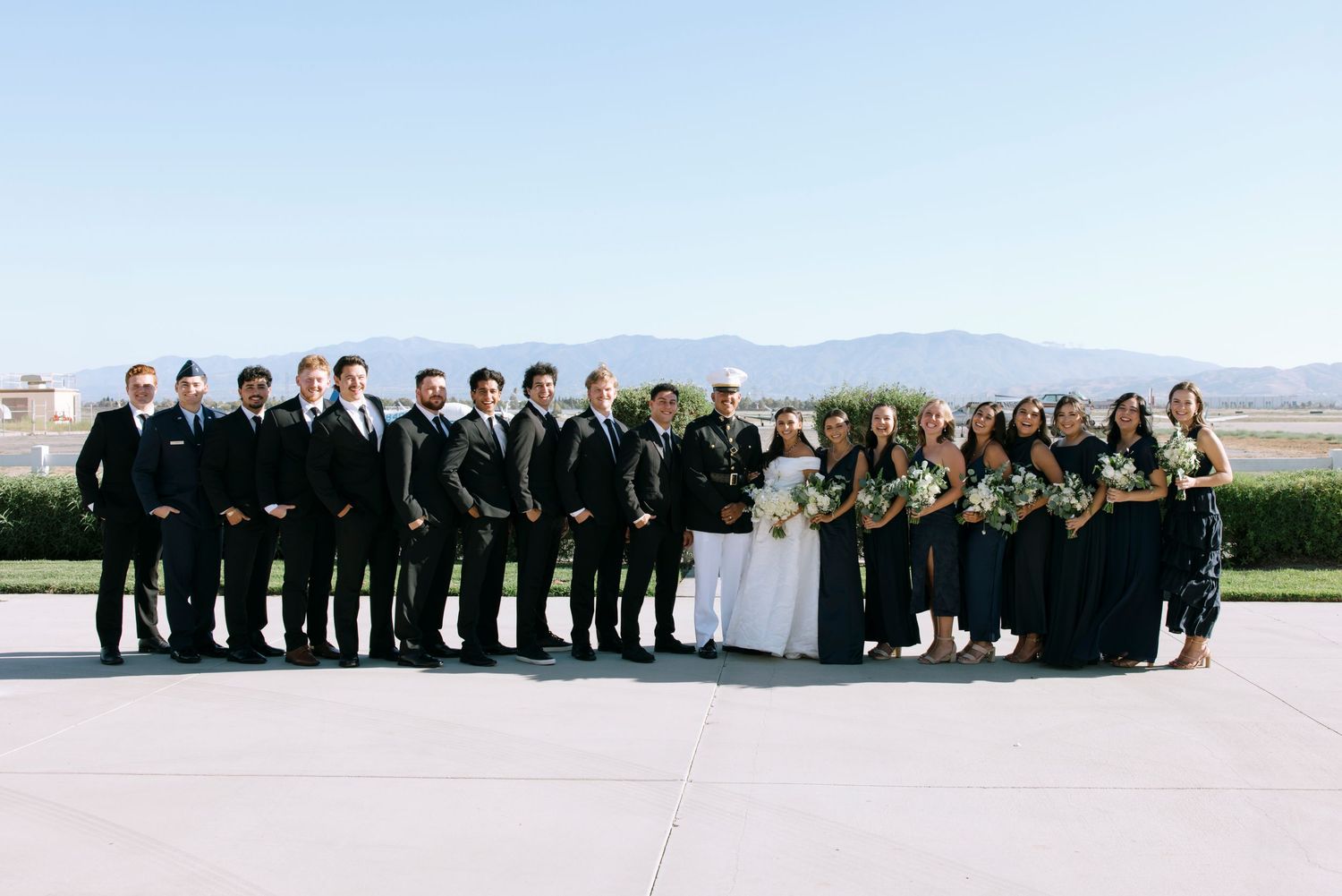 Wedding party members in black and olive green attire pose together outdoors.