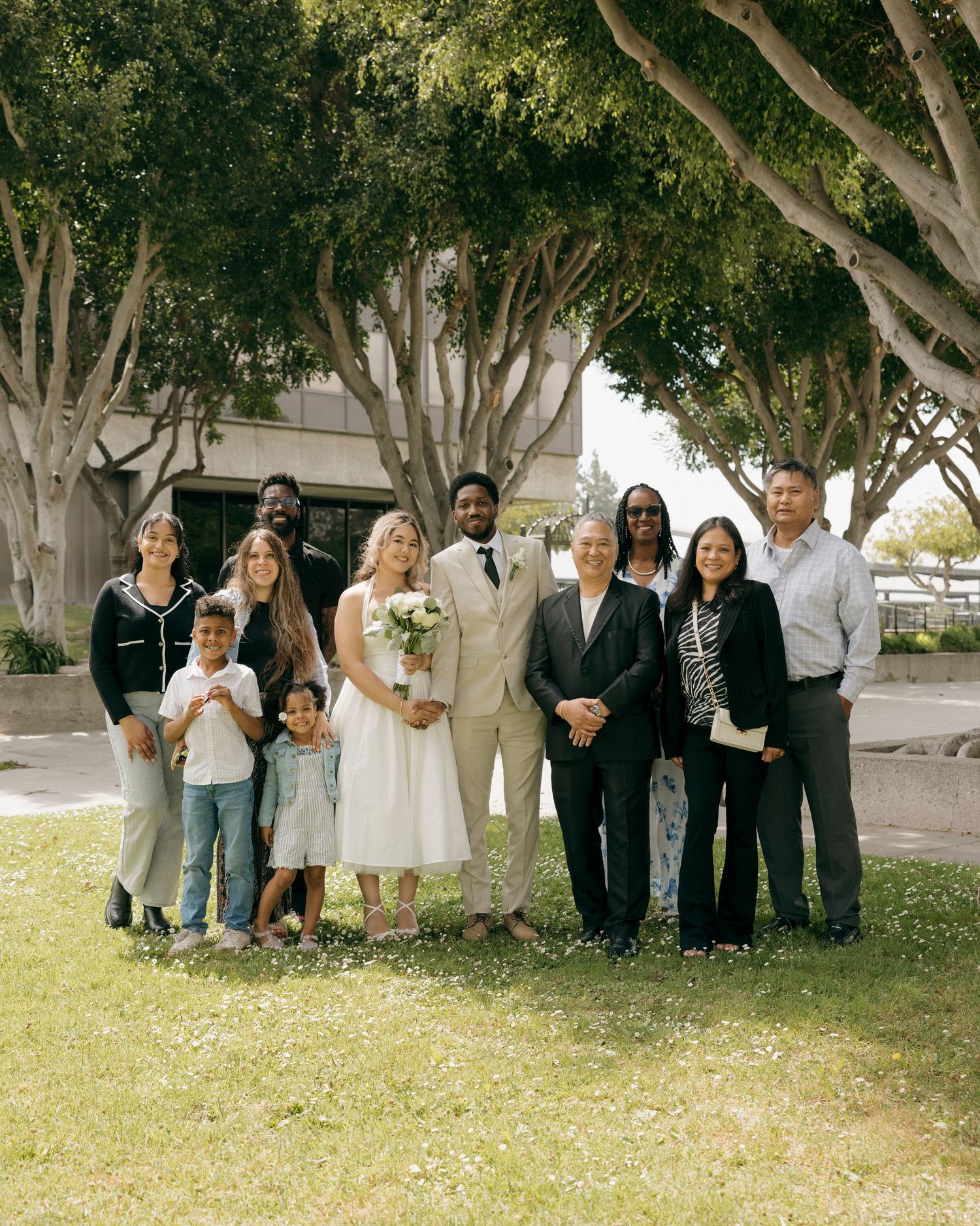 A large group poses together under shady trees in a park setting for a casual outdoor photo.