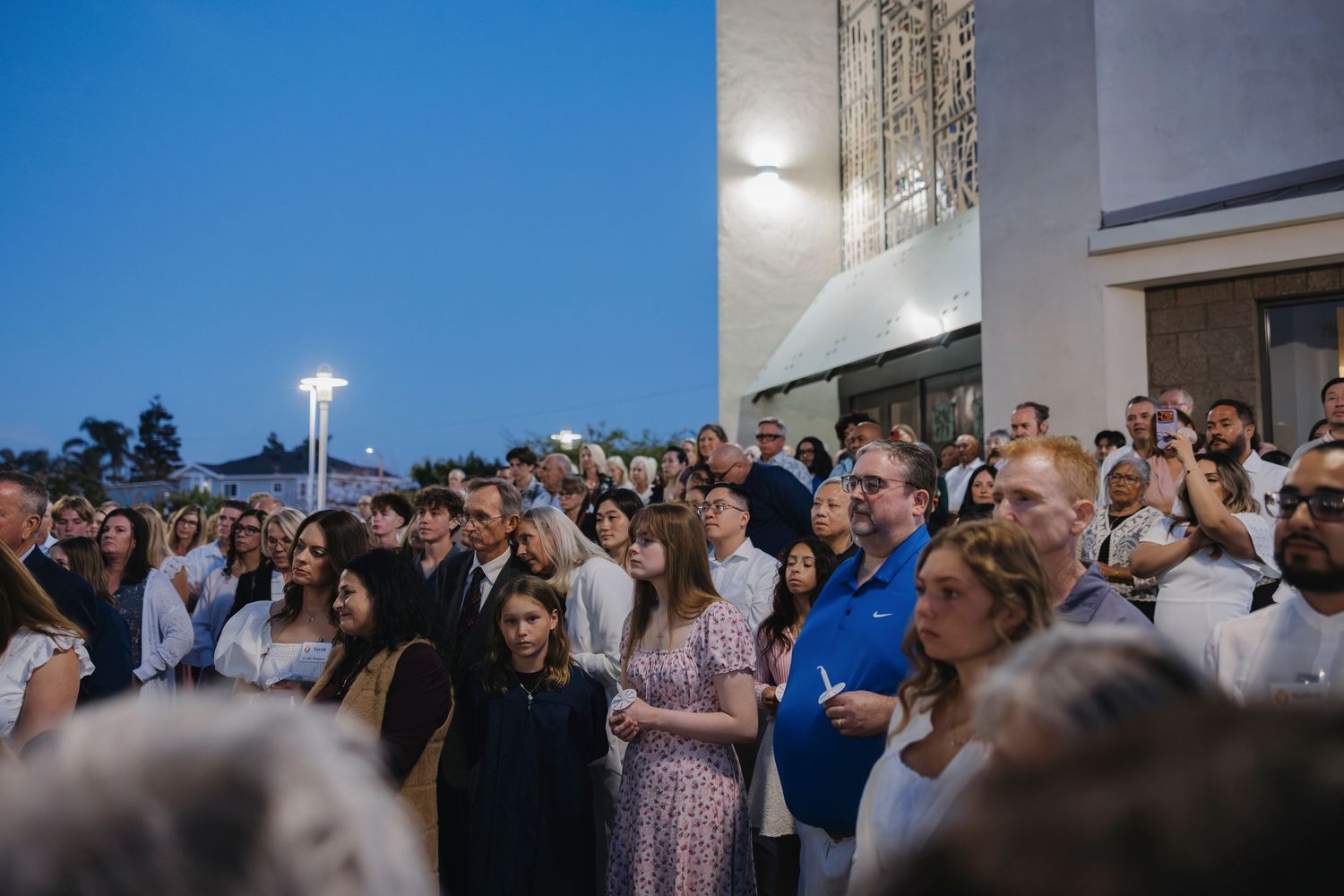 Large crowd gathered outdoors at dusk for a religious ceremony near a modern church building.