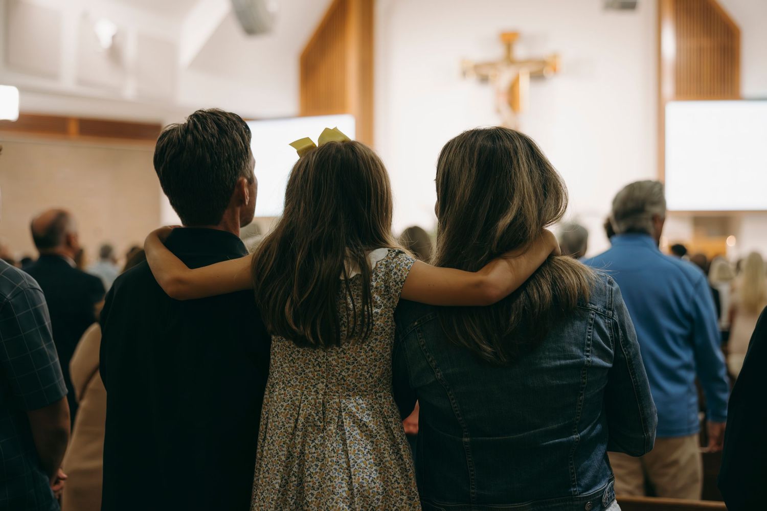 Three people embrace with arms around each other inside a church with a wooden cross on the wall.