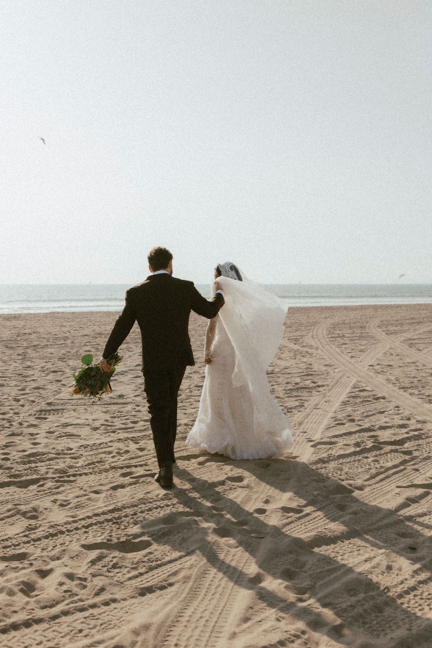 Newlyweds walking hand in hand along a sandy beach leaving footprints behind.