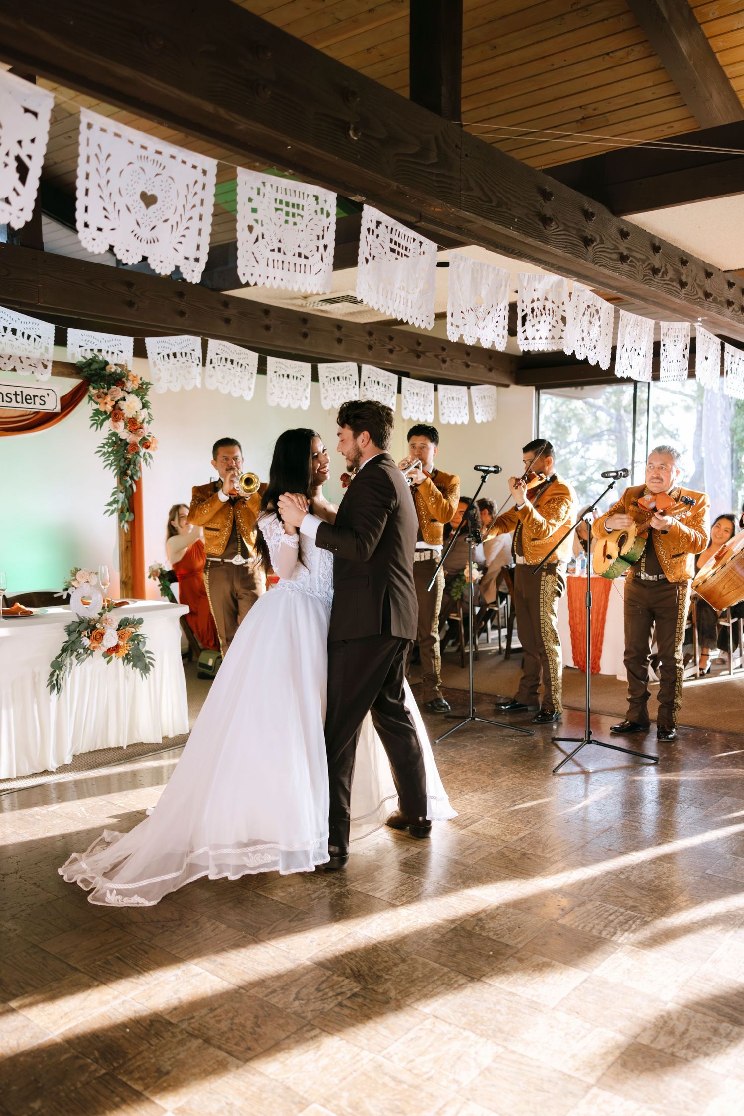 Wedding party dancing in a rustic venue with wooden beams and decorative white papel picado banners.