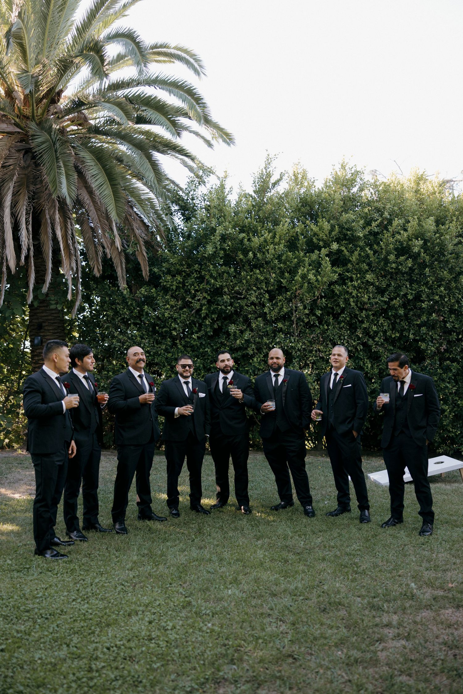 Groomsmen in black suits stand in a line on a grassy lawn with palm trees in the background.