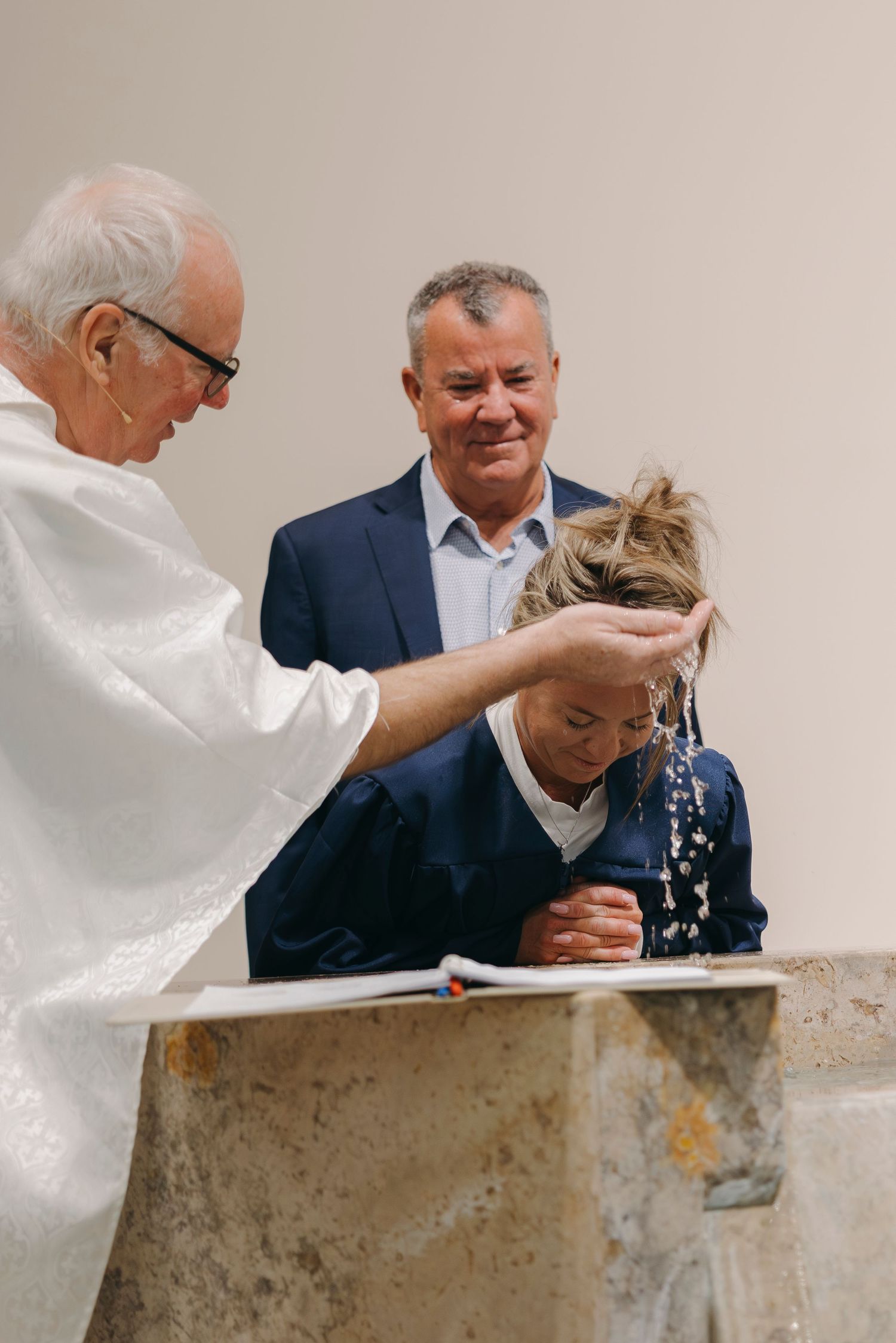 Elderly Catholic priest pouring baptismal water over young girl's head at stone font during baptism ceremony.
