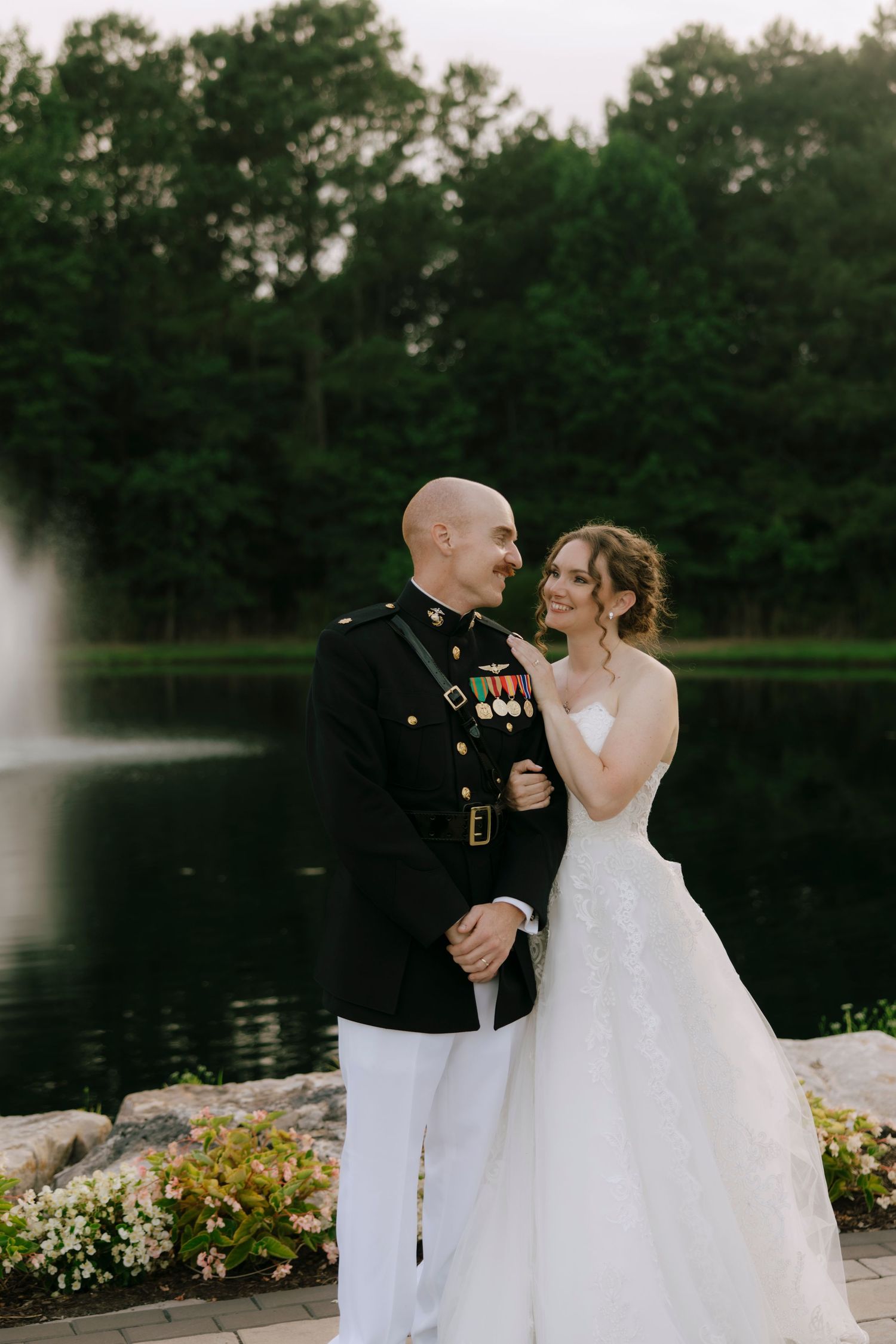 Military groom in dress uniform gazes at bride by a serene pond with fountain backdrop.