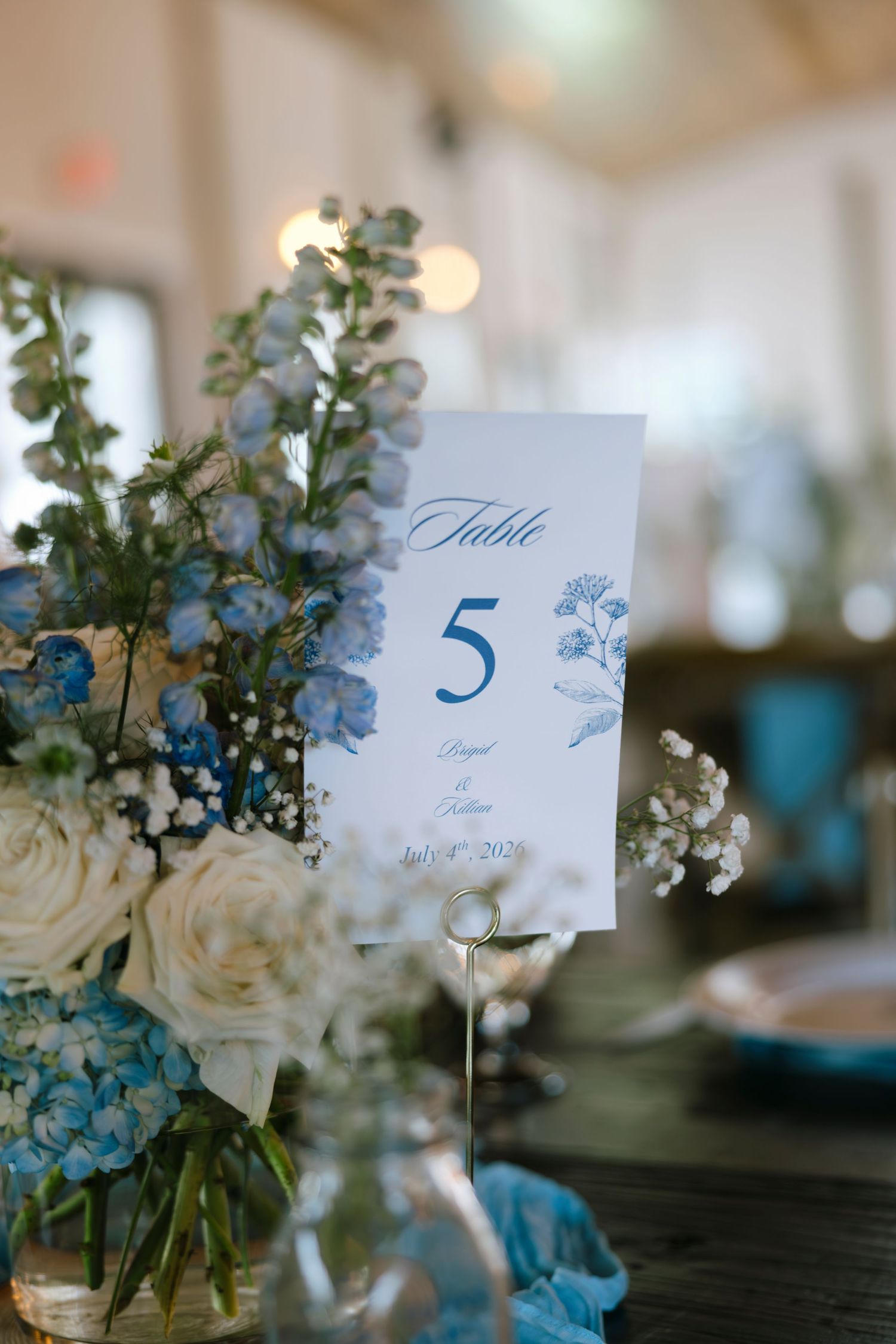 Blue and white wedding table number 5 card with floral centerpiece featuring hydrangeas and roses dated July 4th 2026.