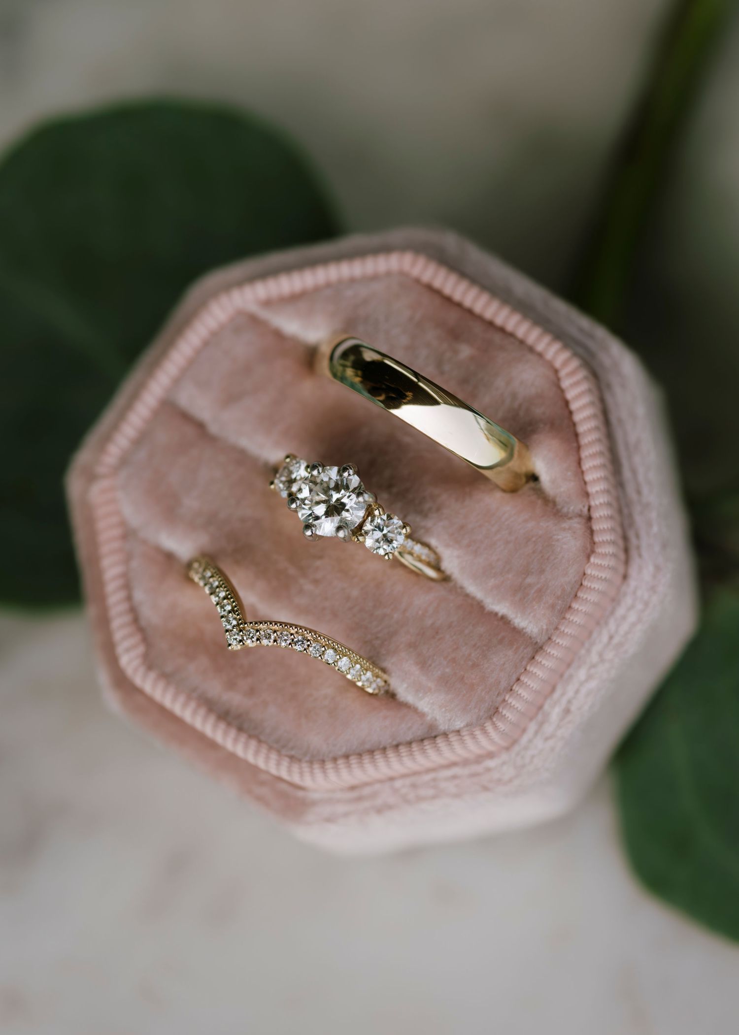 Three gold rings displayed in a pink velvet hexagonal ring box, featuring a diamond cluster engagement ring, chevron band, and plain band.
