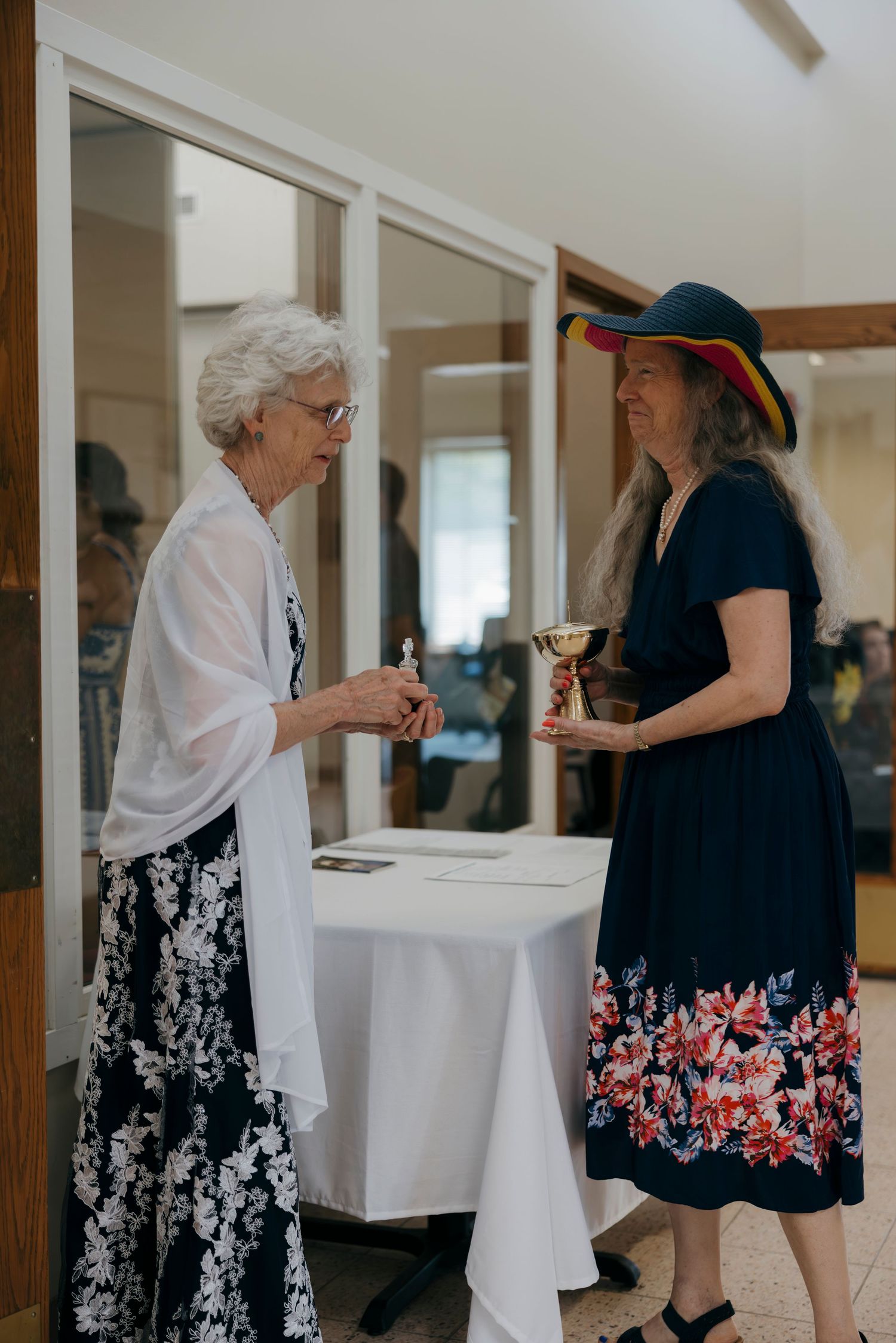 Two elderly women in elegant dresses chat at a white table, one wearing a colorful hat, at a formal indoor event.