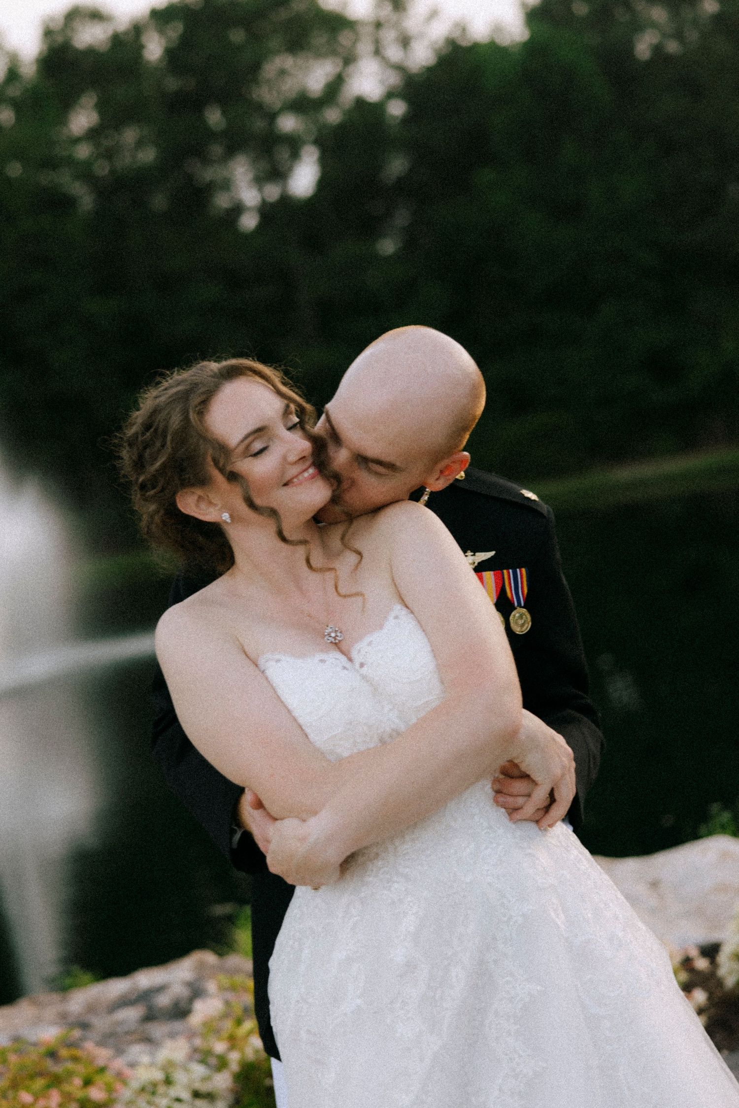 A groom in military dress uniform kisses his bride in a white lace gown outdoors near a pond.