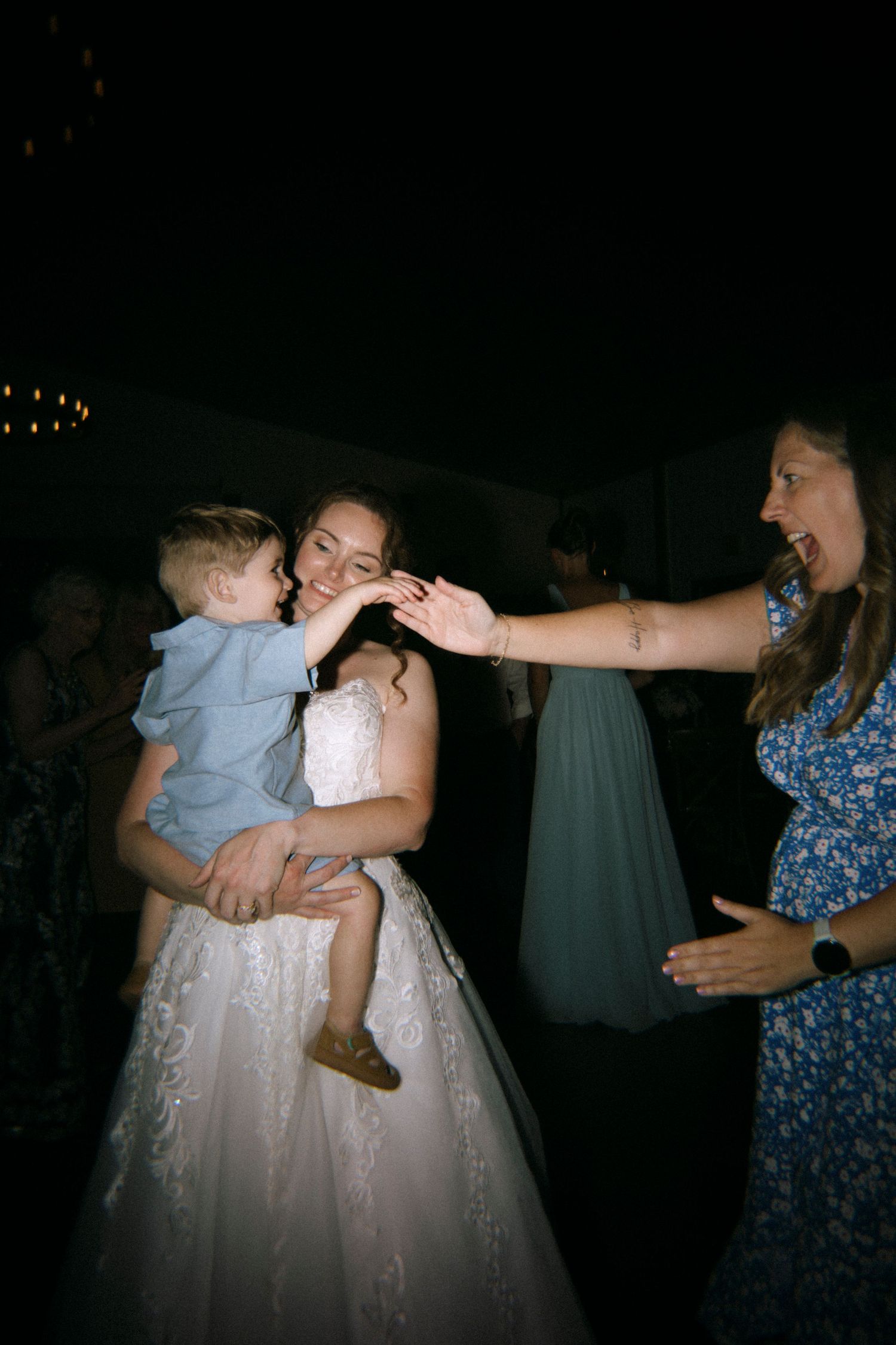Bride in white gown holds baby at wedding reception while guest reaches out to interact with the child.