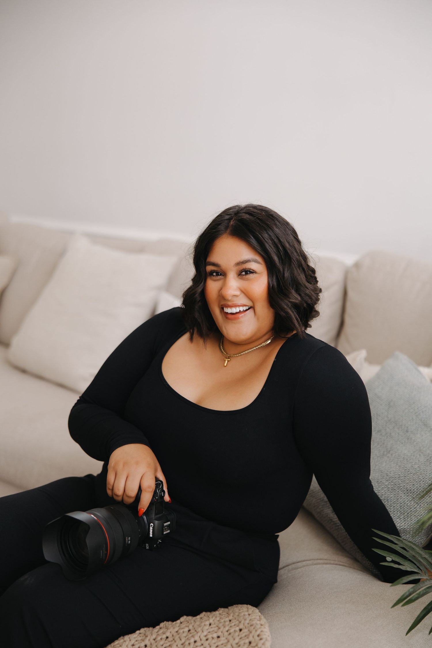 Professional photographer in black attire sitting on beige couch with camera equipment.