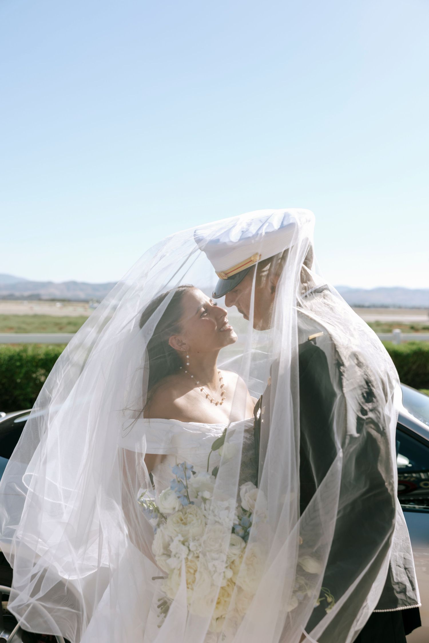Wedding couple embracing under a delicate white veil with mountains in the background.