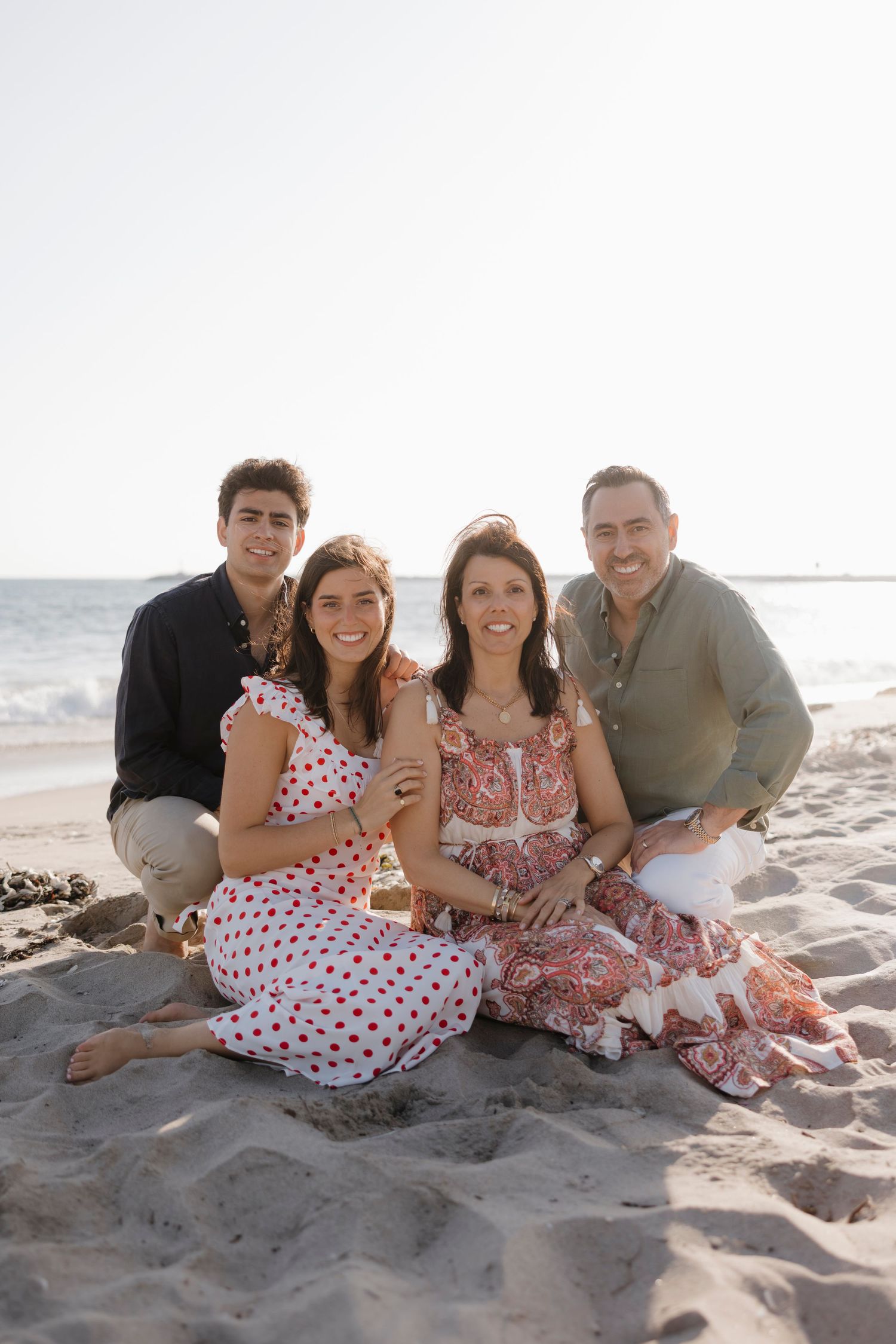 Family of four smiling together on a sandy beach, with ocean waves in the background on a sunny day.