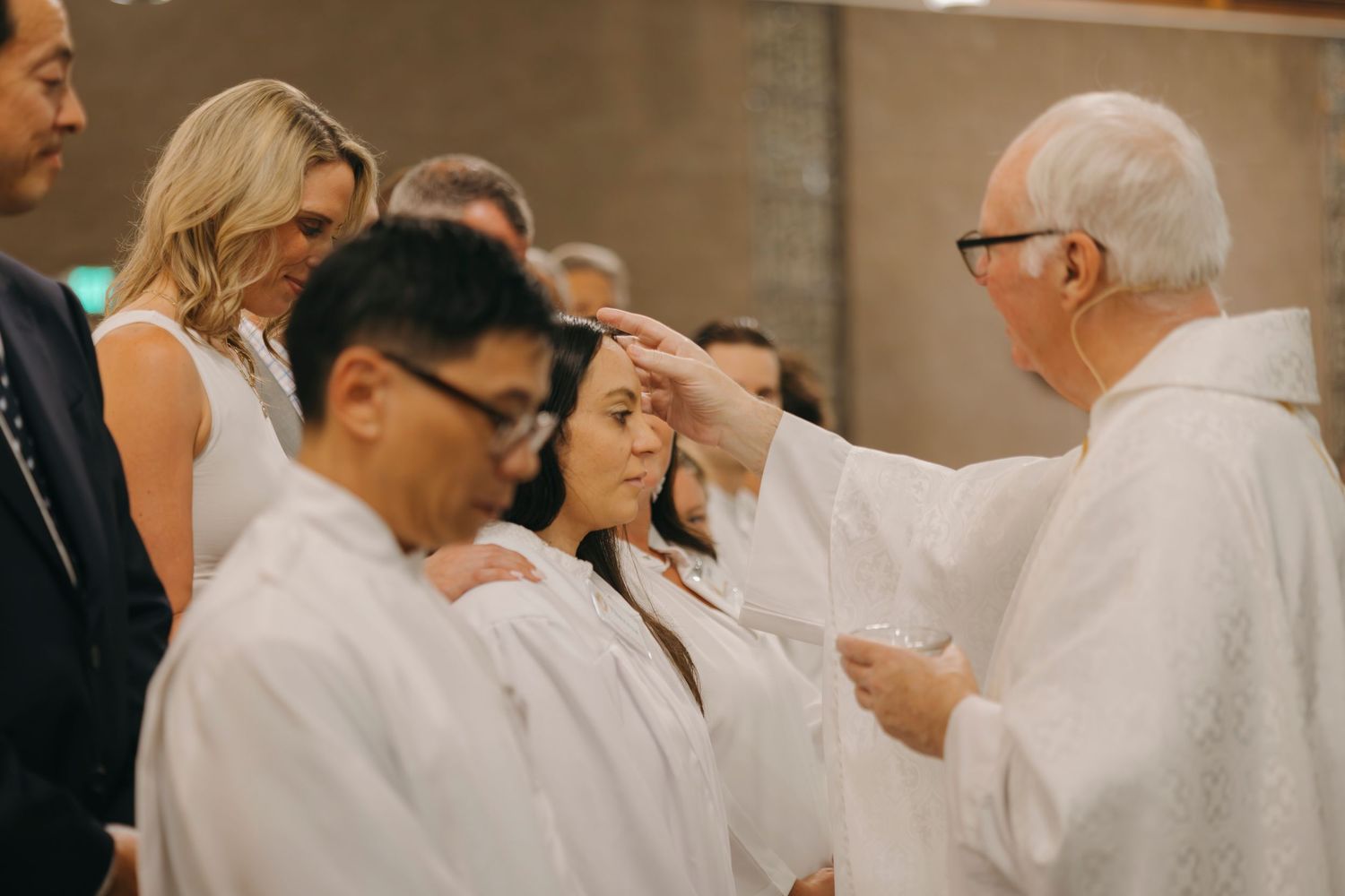 Priest performing confirmation anointing on woman in white robe during Catholic religious ceremony with congregation gathered.
