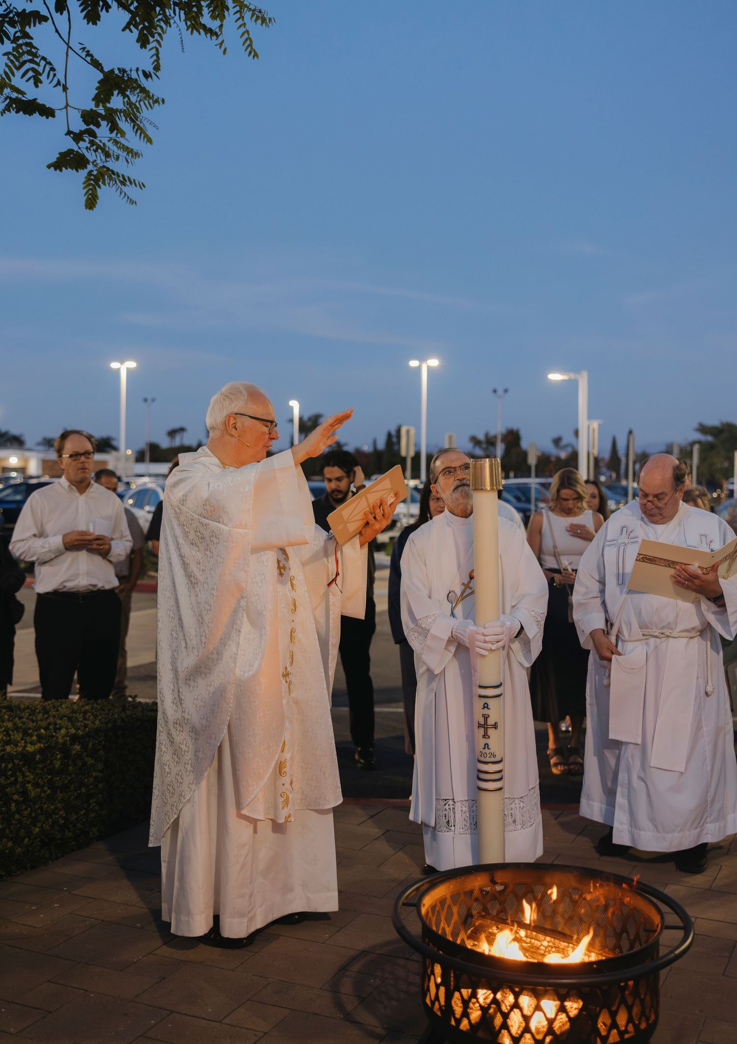 Catholic priest blessing Easter fire at dusk outdoor ceremony with clergy holding Paschal candle.
