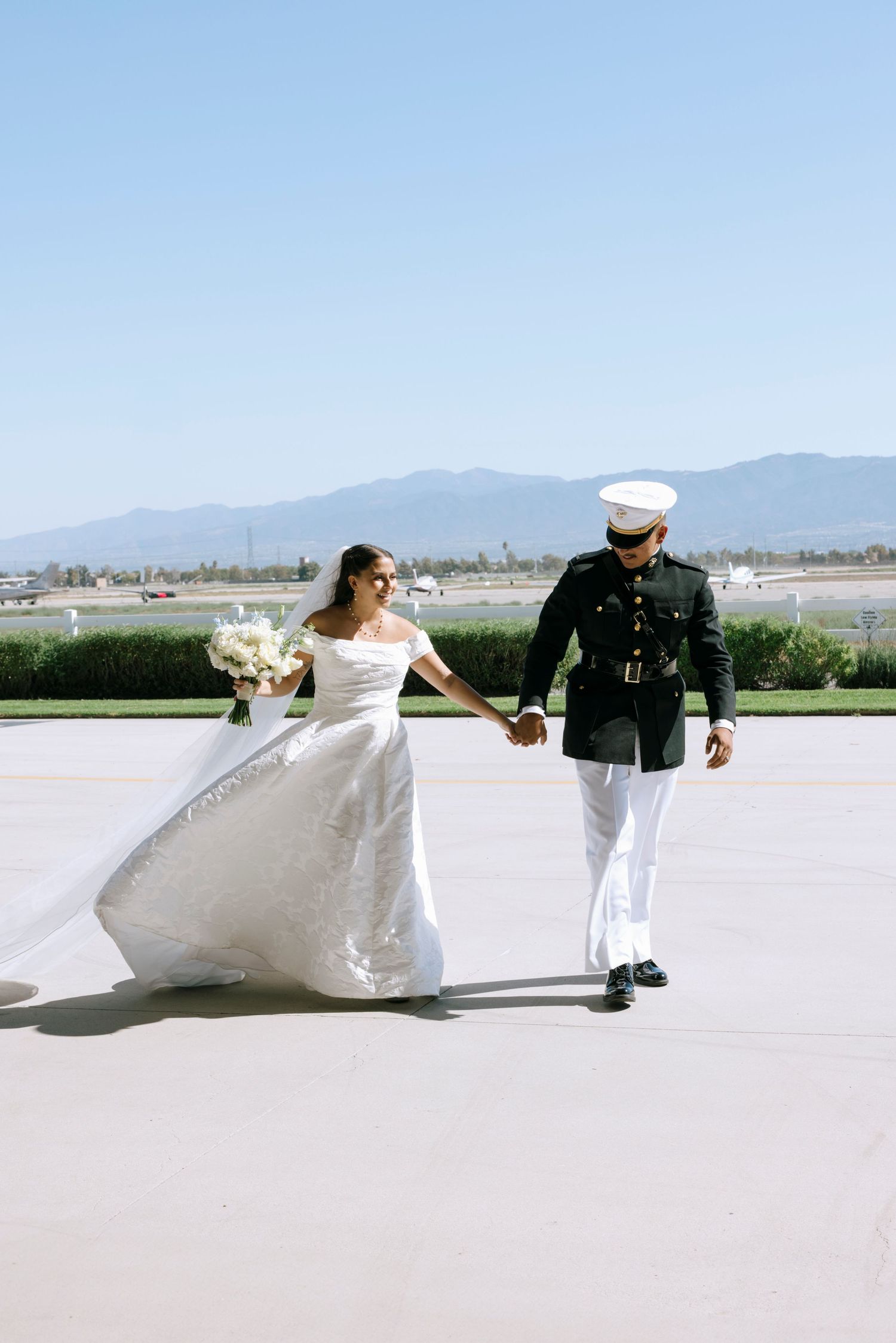 A couple in wedding attire walks together against a mountain backdrop on a bright day.