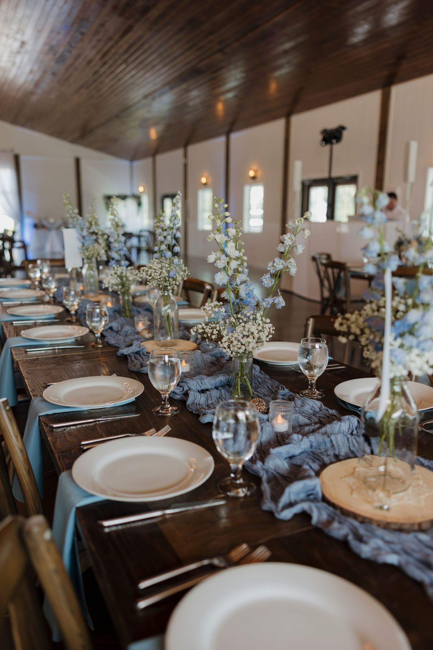 Elegant wedding reception table with blue runner, white plates, crystal glasses, and delicate floral centerpieces in a rustic venue.