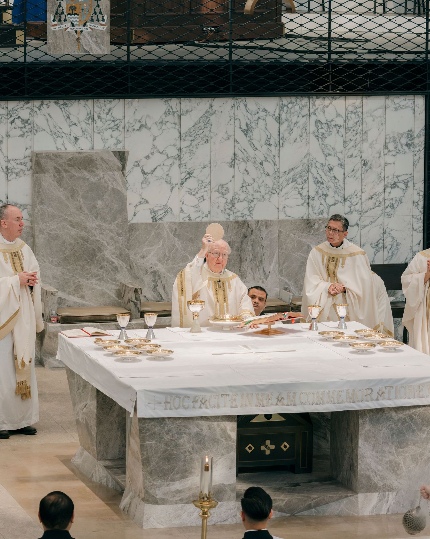 Wide view of clergy members in white robes seated at long table against marble wall.
