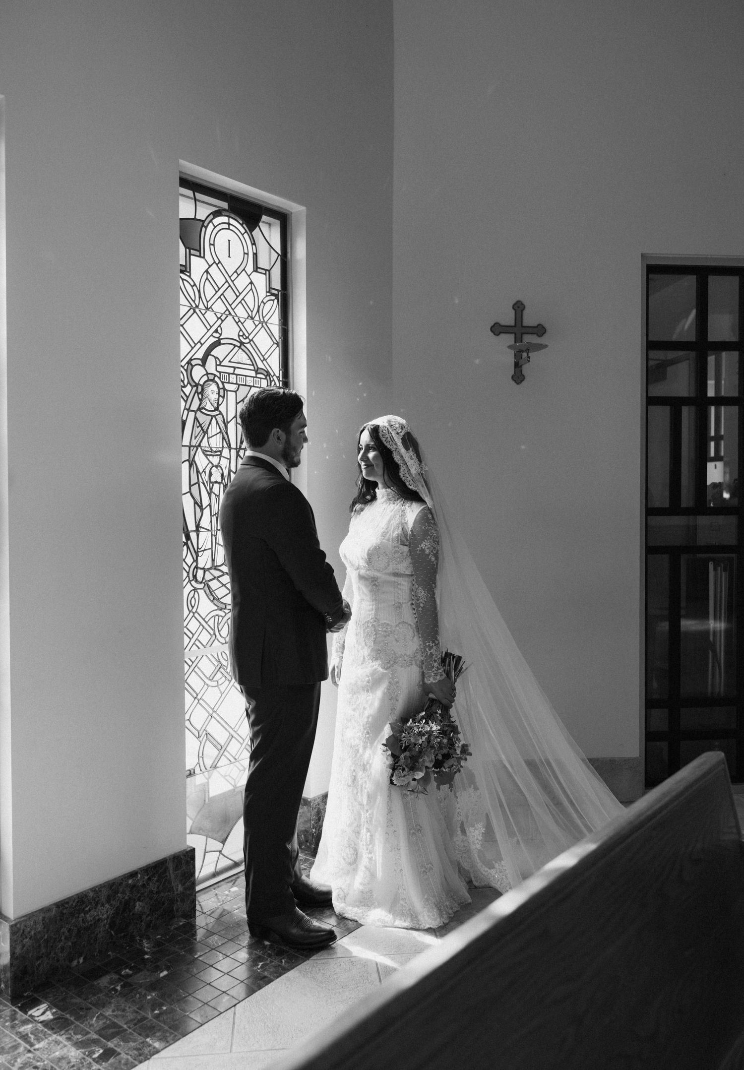 A couple in wedding attire stands by a stained glass window in a church.