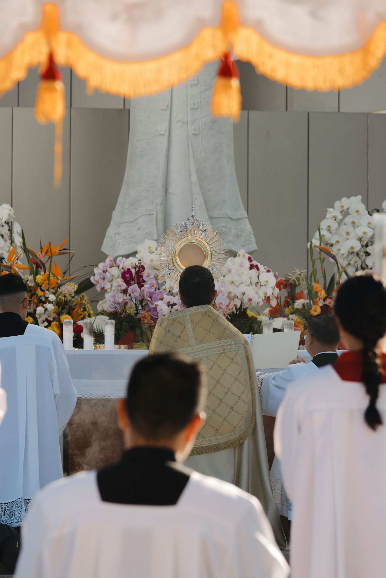 Congregation in white robes attends outdoor Eucharistic celebration with orange banners and floral decorations.