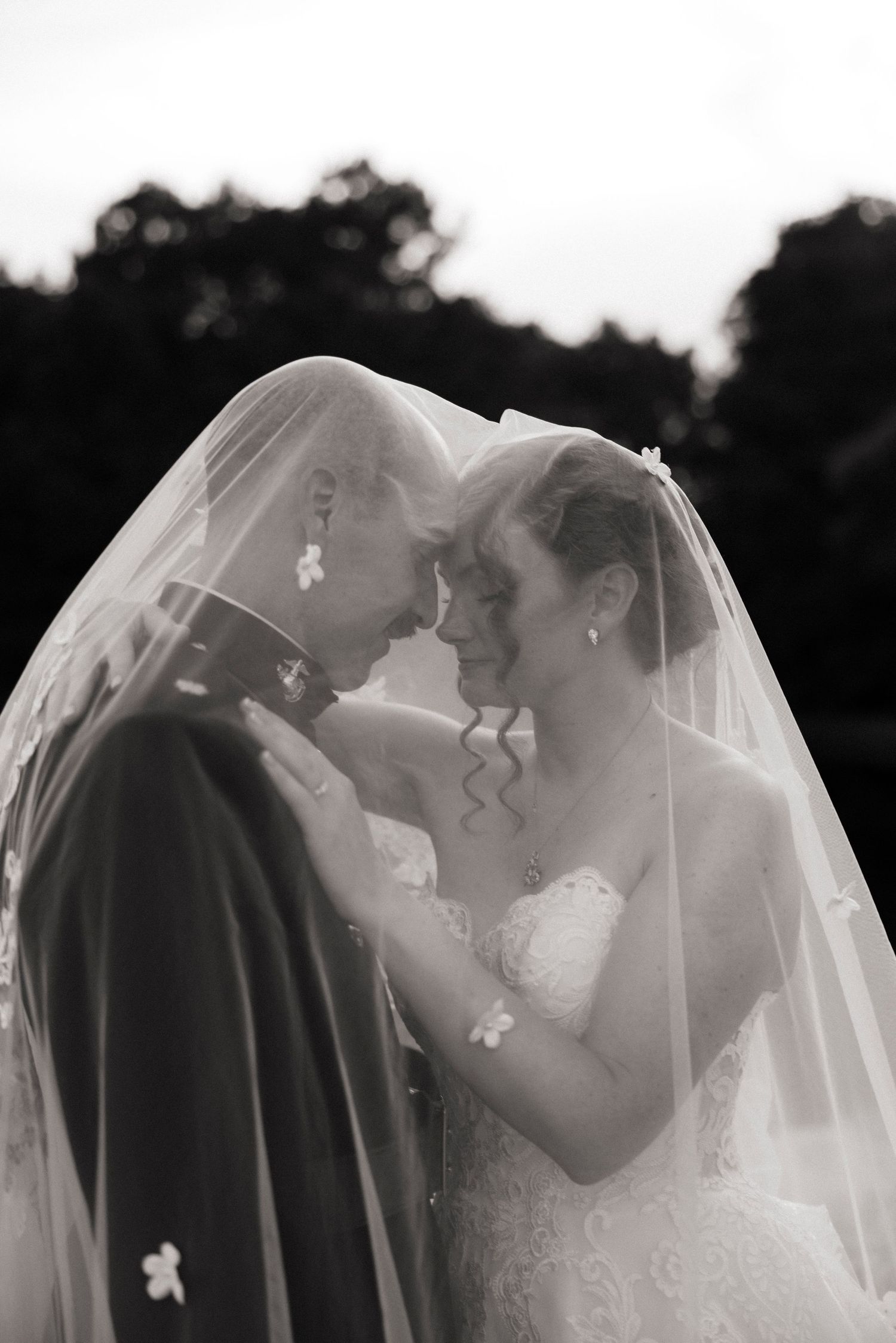 Black and white photo of bride and groom sharing intimate moment under wedding veil, foreheads touching, smiling.