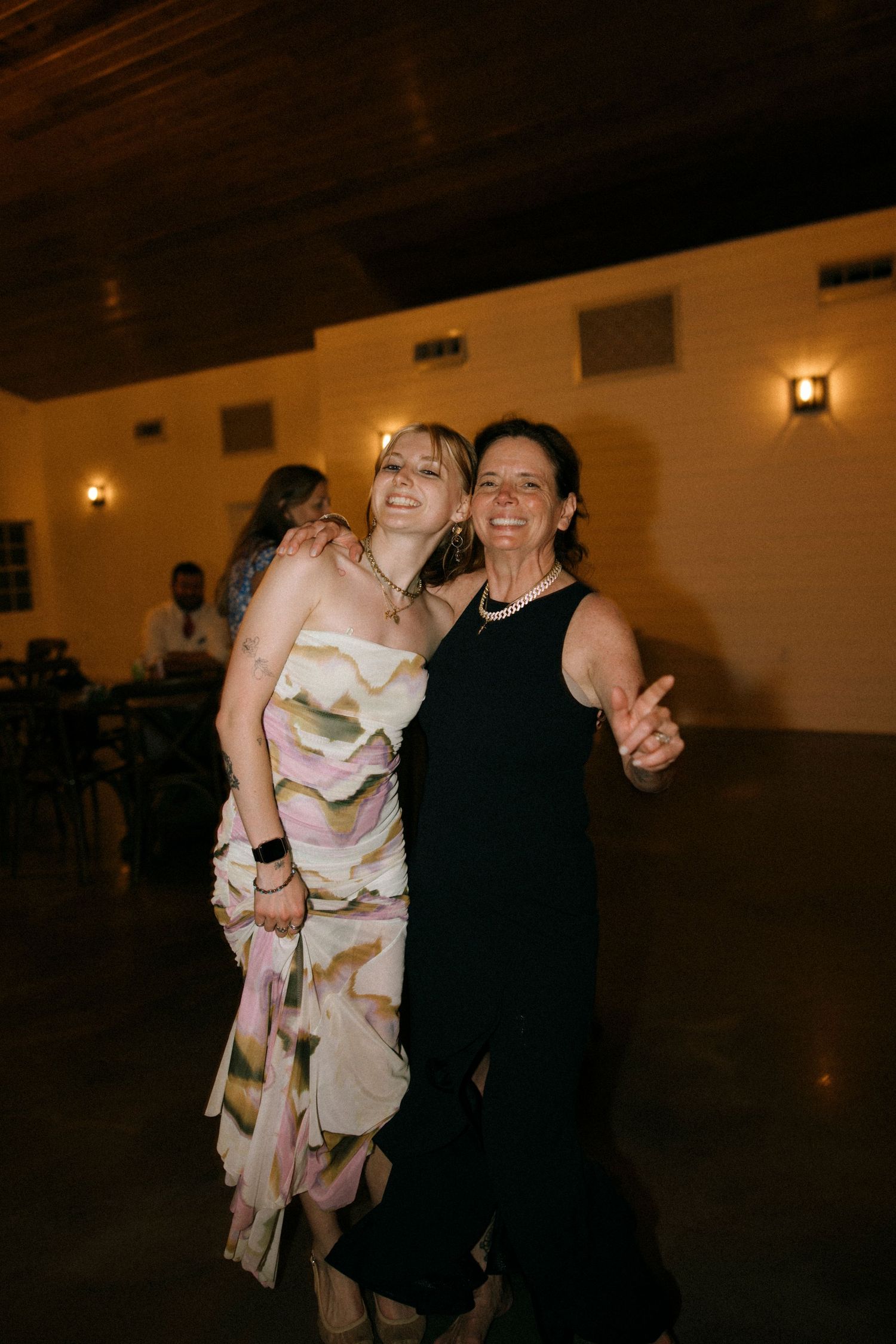 Two women smiling and posing together at a wedding reception, one in a floral dress and one in a black gown.