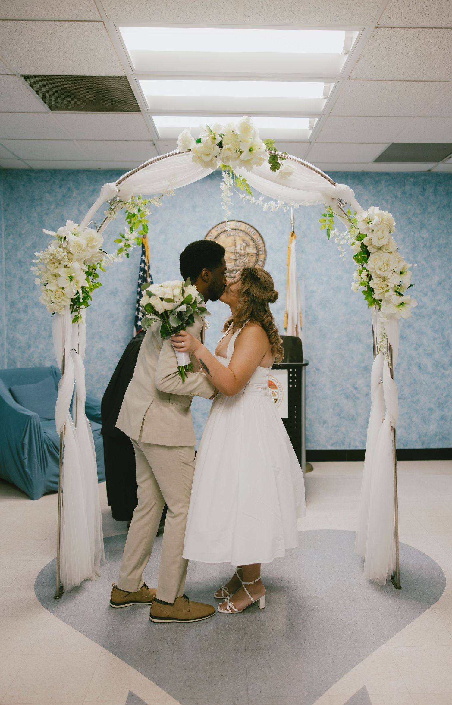 Newlyweds share their first kiss under a white floral arch decorated with orchids during an indoor ceremony.