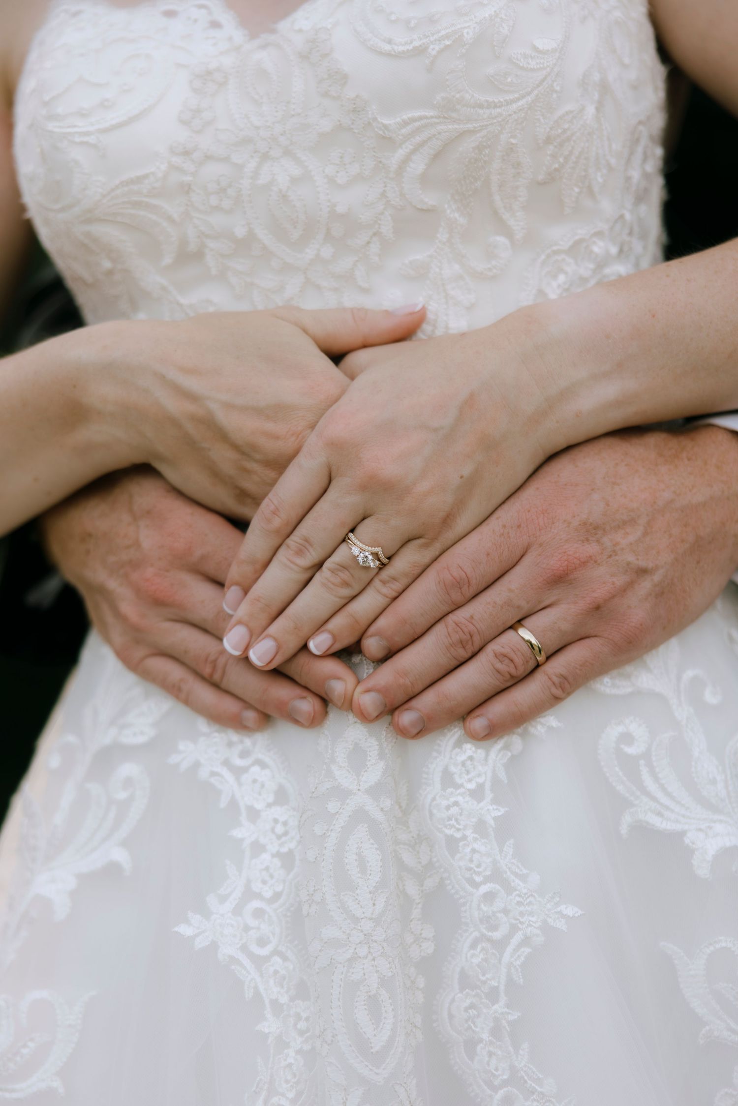 Bride and groom displaying wedding rings on hands clasped together over white lace bridal gown.