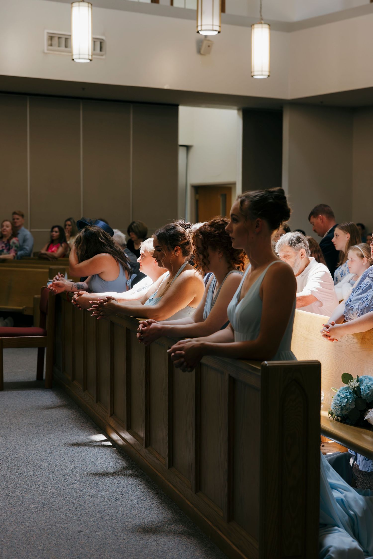 Wedding guests in church pews bow heads in prayer during ceremony, bridesmaids in blue dresses visible.
