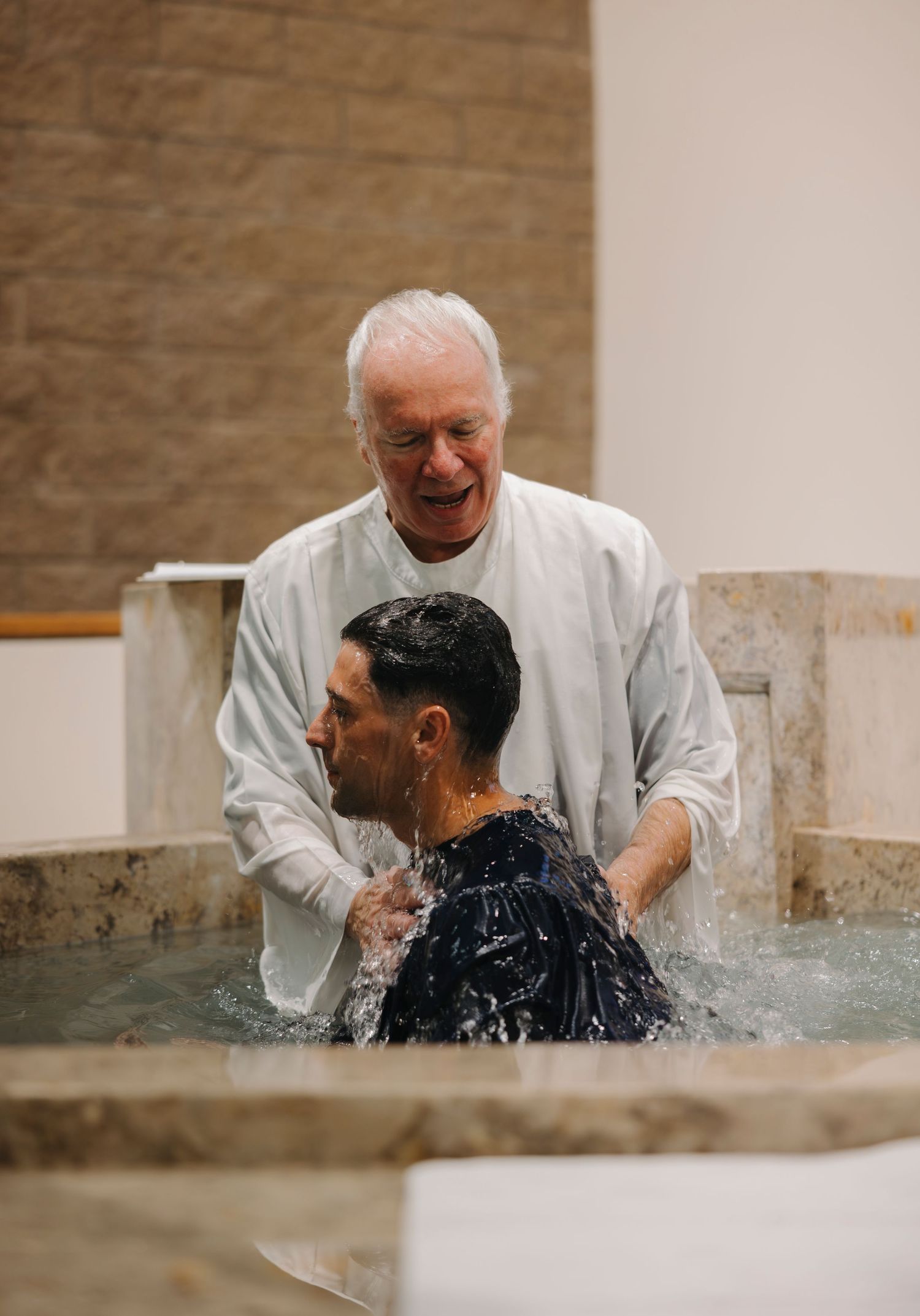 Elderly pastor smiling joyfully while baptizing a man in a church baptismal pool.