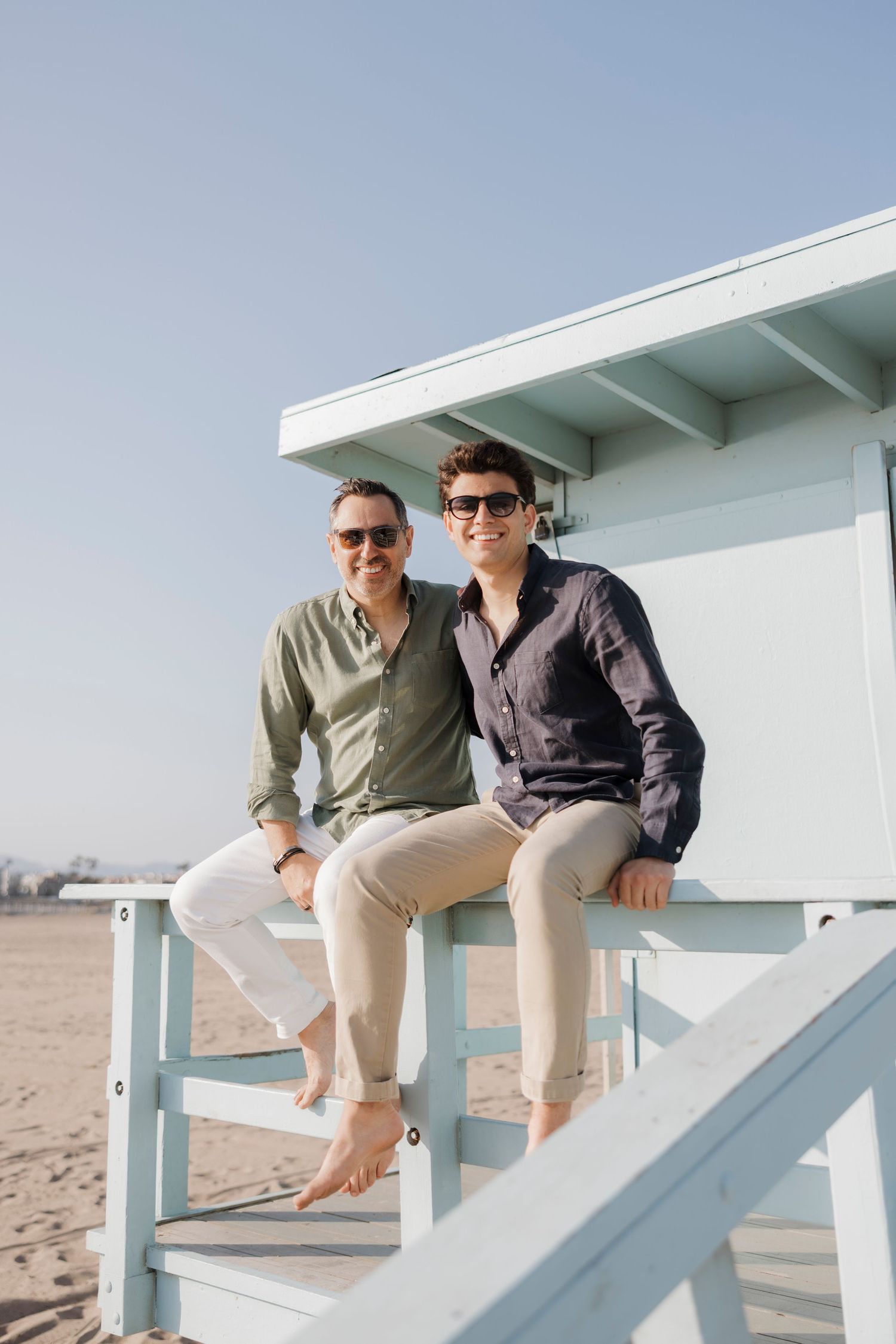 Two men sitting together on a lifeguard tower at the beach on a sunny day.