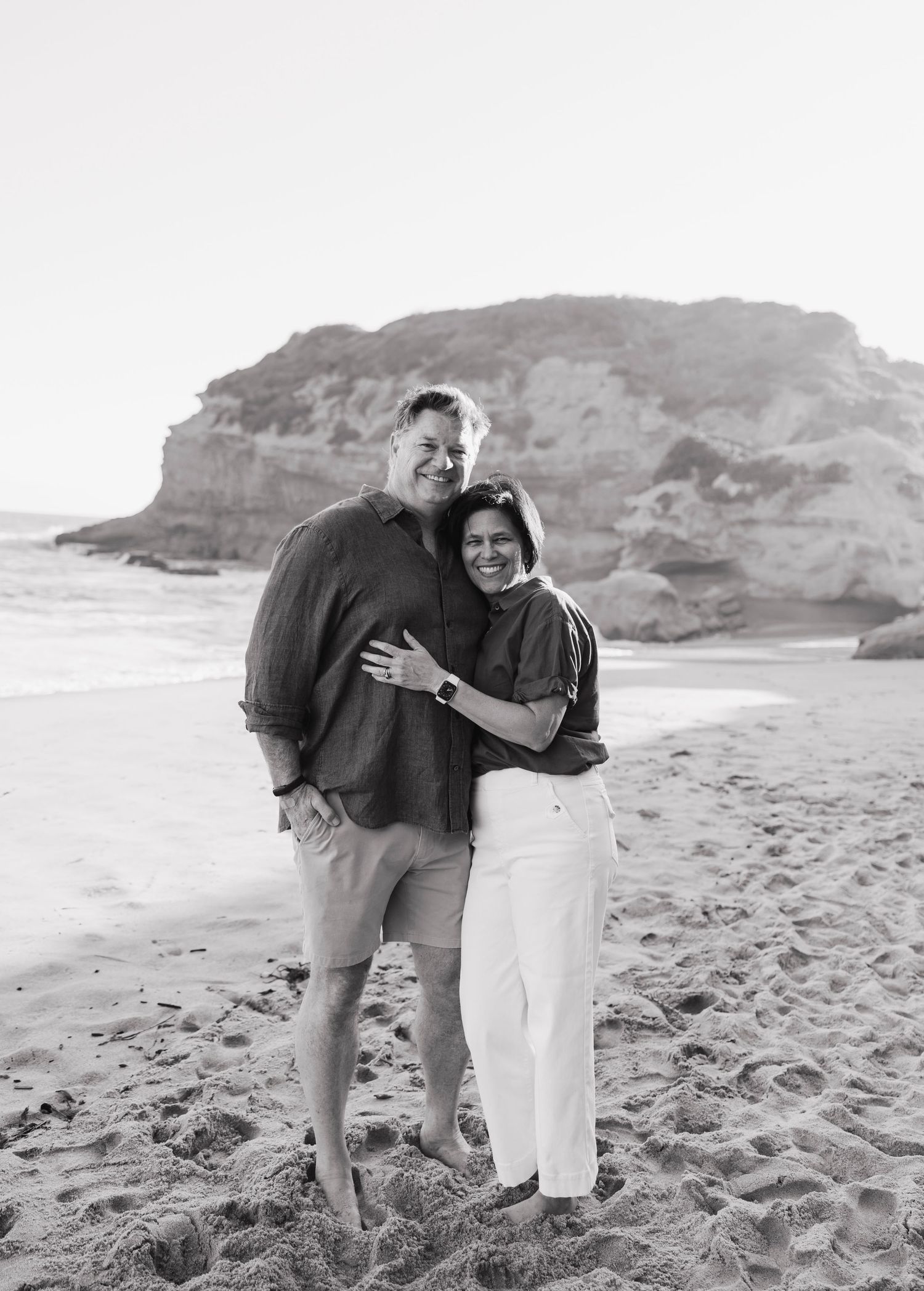 Black and white photo of a smiling couple embracing on a sandy beach with rocky cliffs in the background.