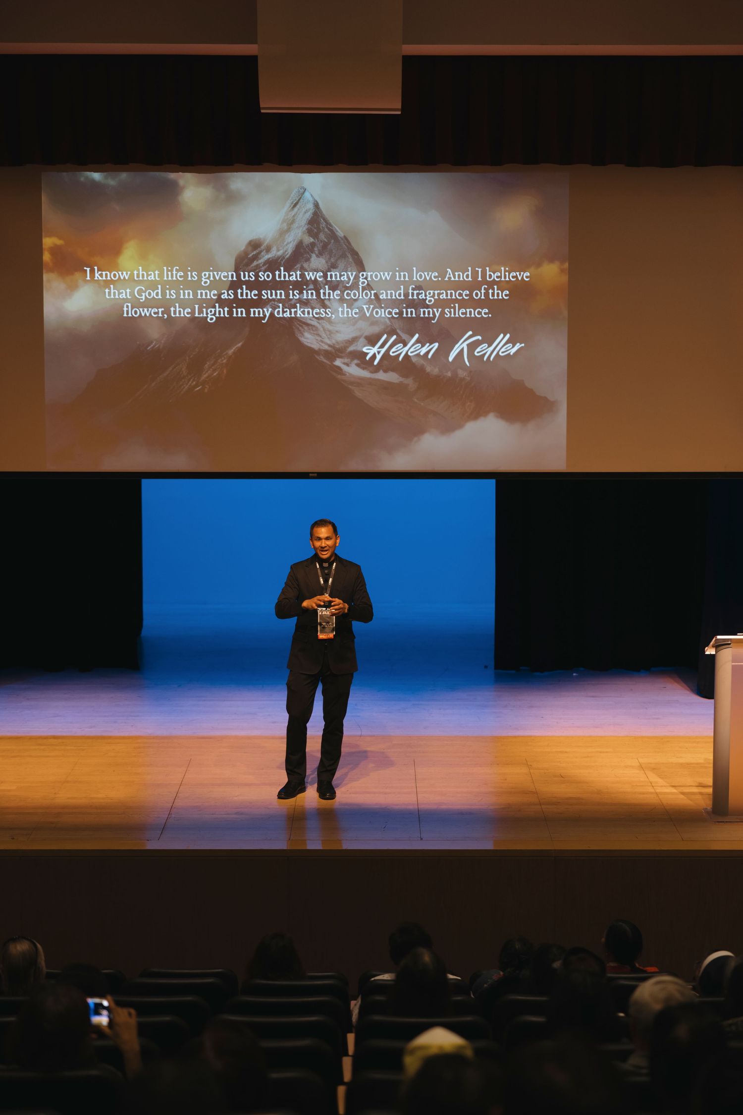 A presenter on stage with an inspirational mountain quote projected on the screen behind.