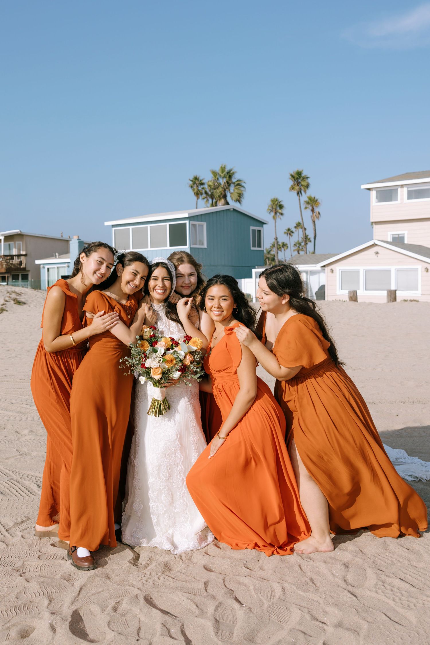 Wedding party in bright orange dresses creates joyful poses on the sand with palm trees in background.