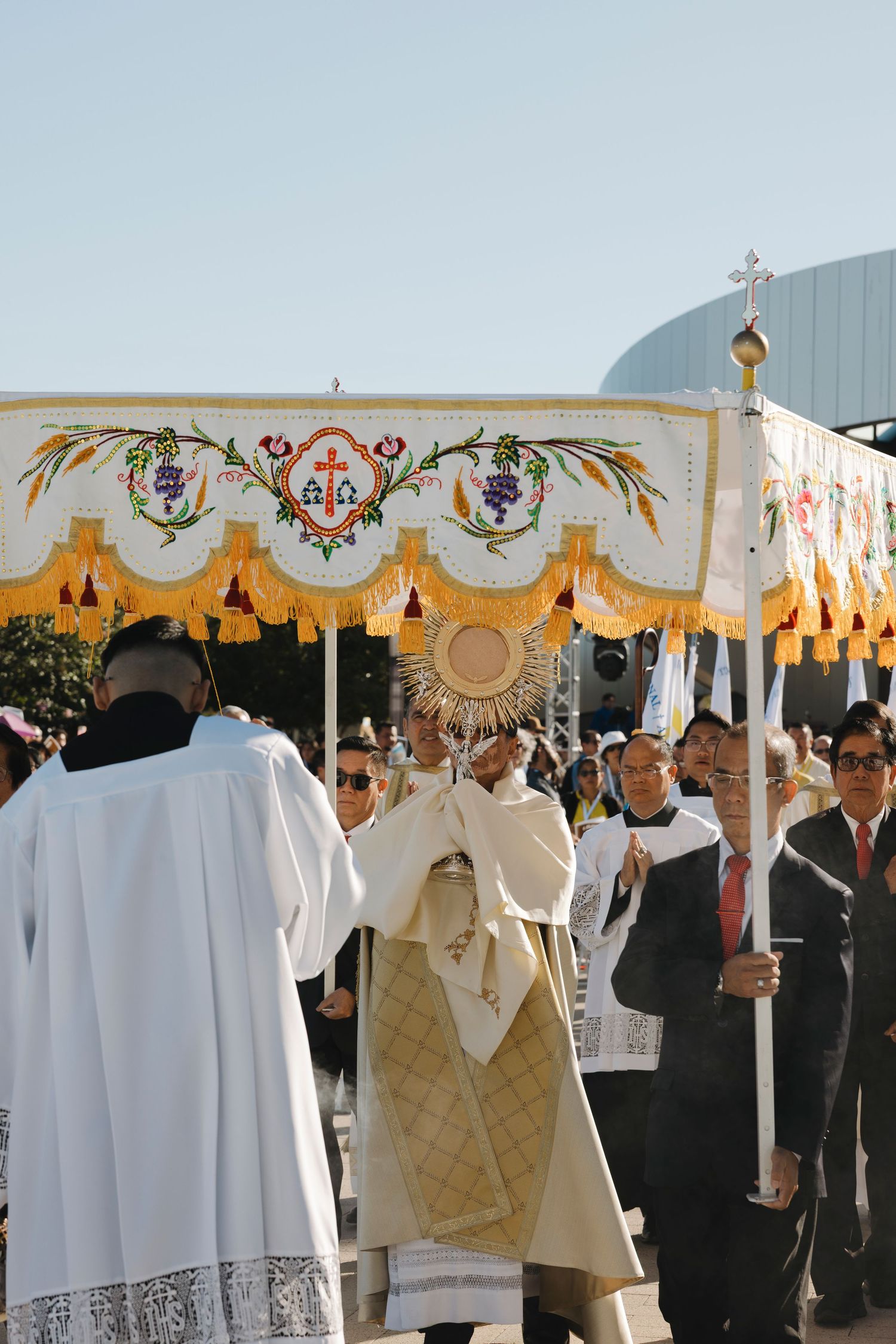 Decorated ceremonial canopy with religious symbols being carried during outdoor Catholic procession.
