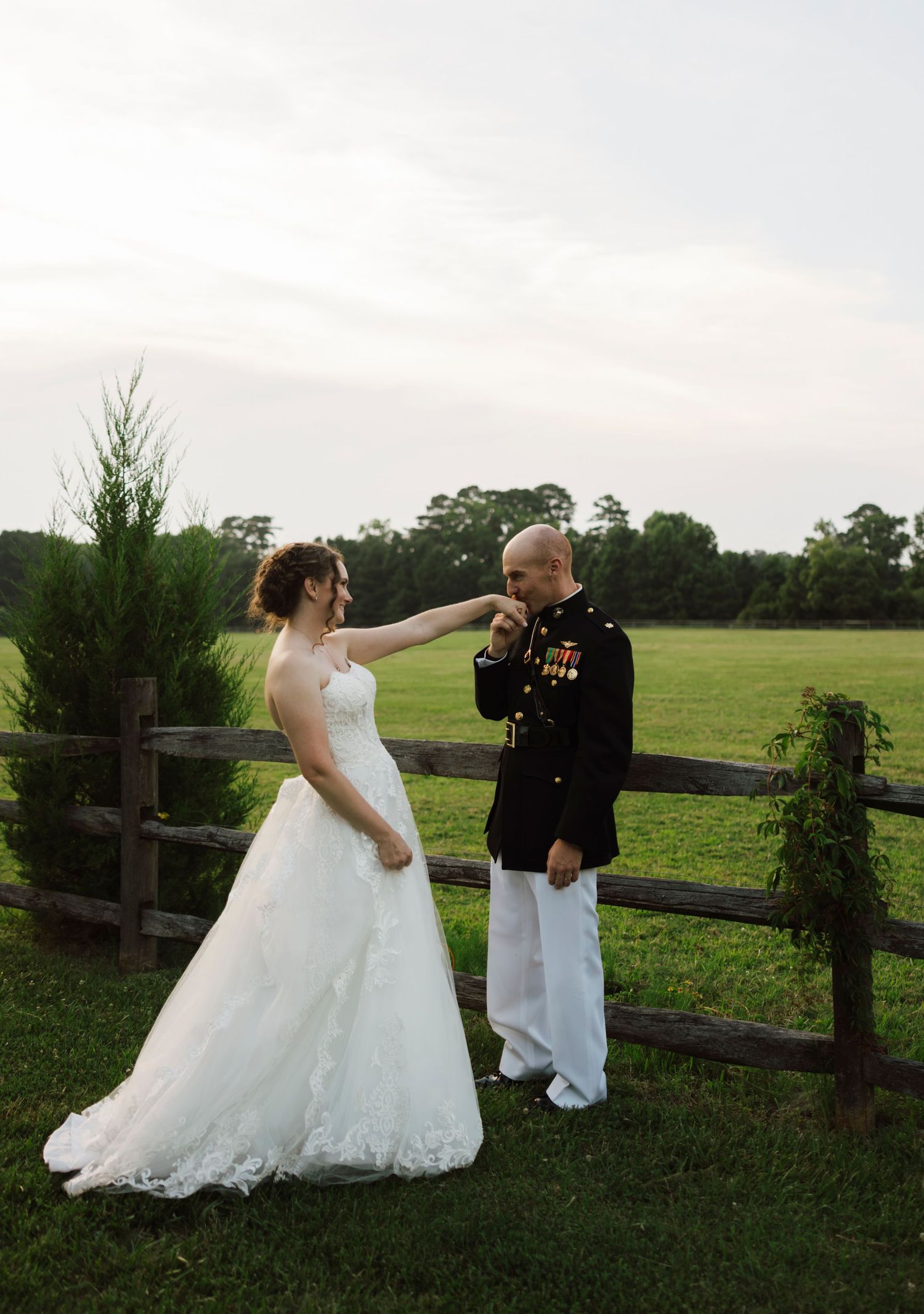 Marine groom kisses bride's hand in dress uniform at outdoor wedding ceremony by rustic fence and green field.