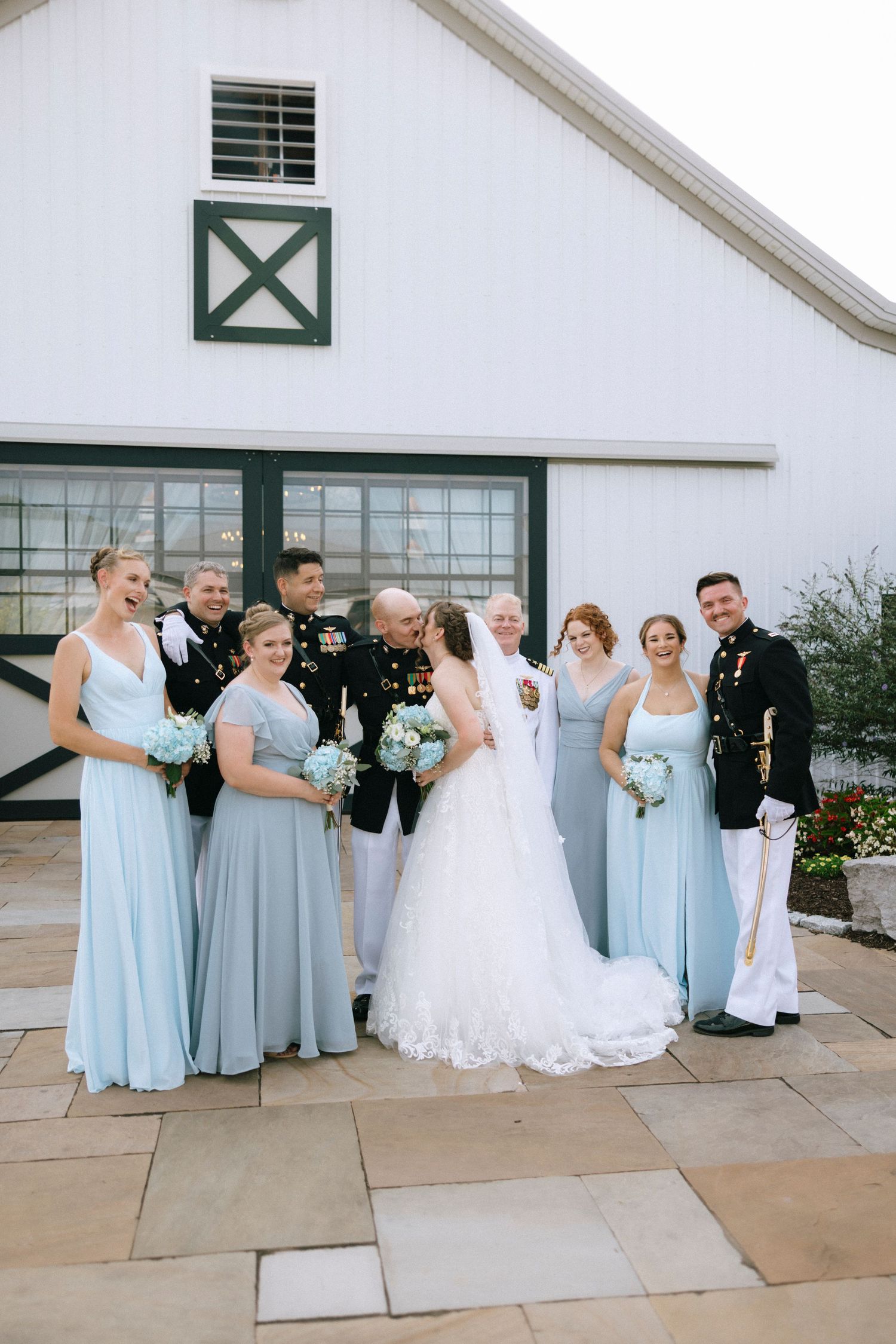 Wedding party poses outside white barn, bridesmaids in light blue gowns with bouquets, groomsmen in military dress uniforms.