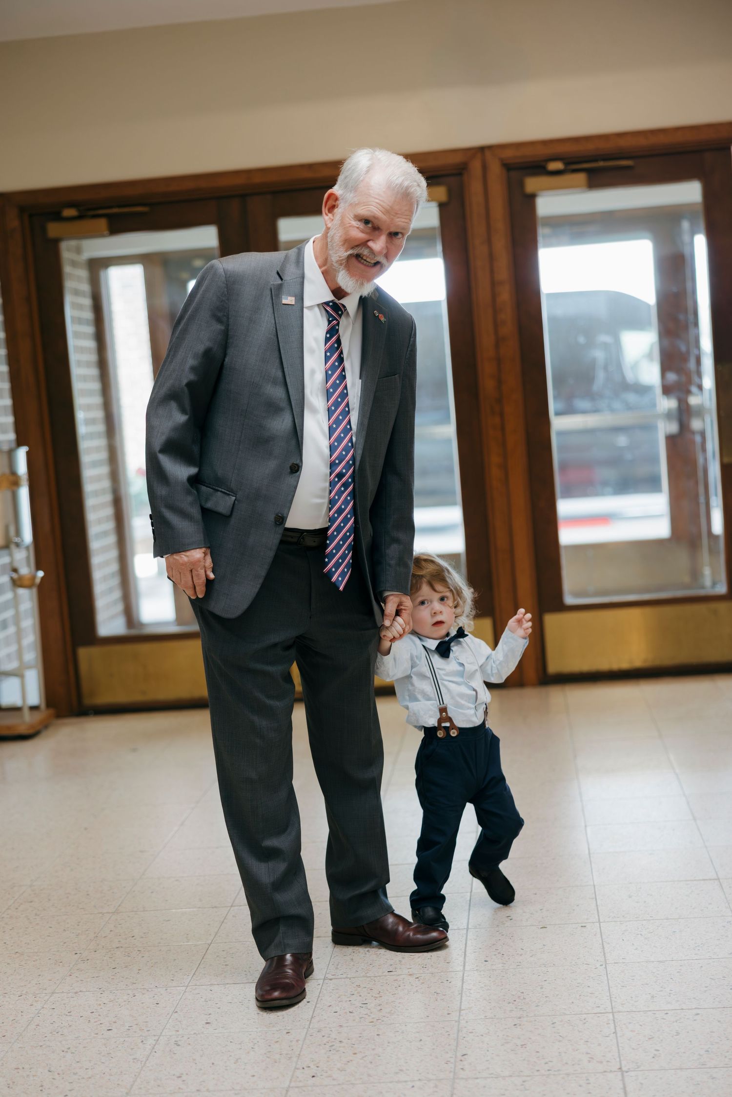 Elderly man in gray suit holds toddler boy's hand while walking together in a formal building.
