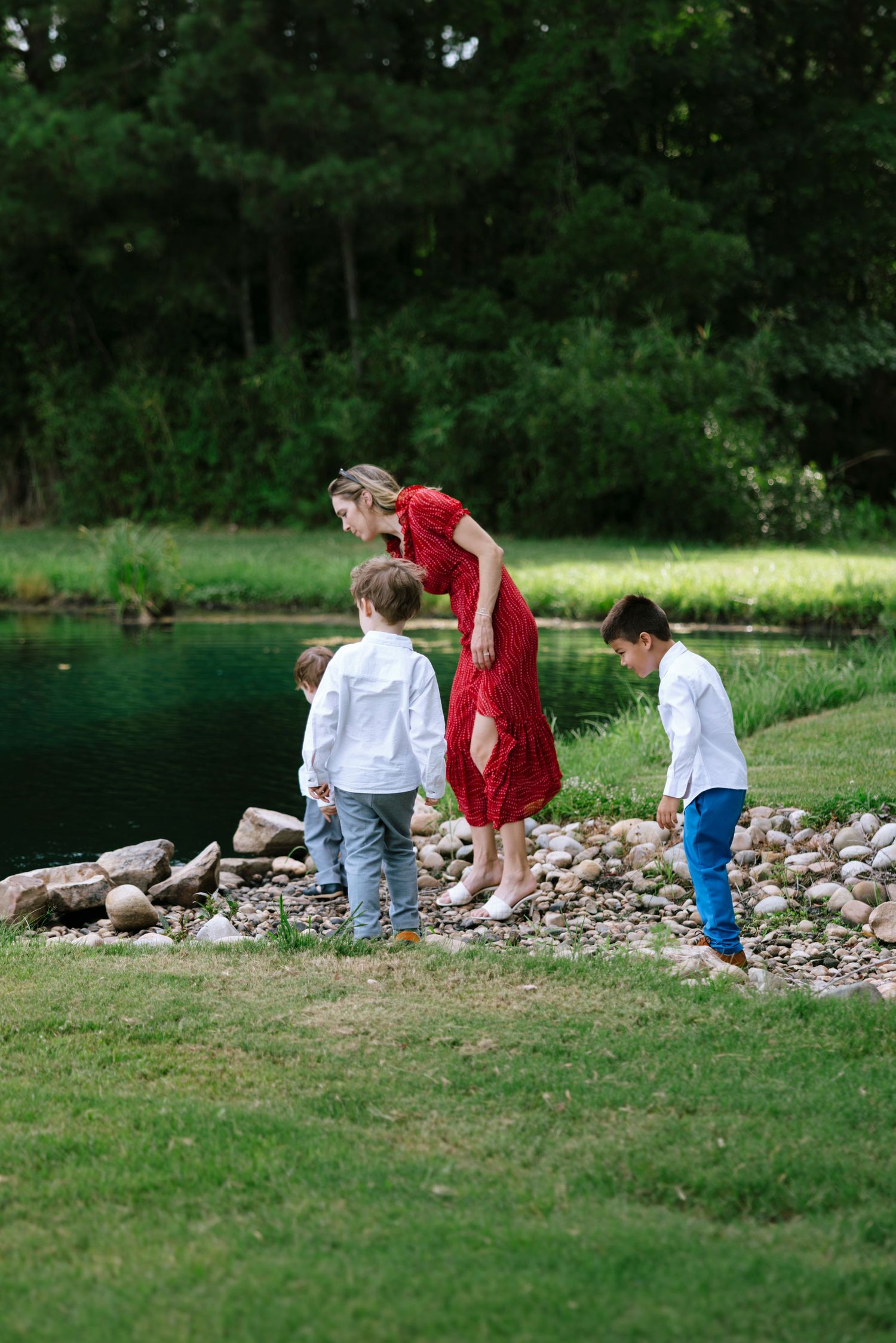 A woman in a red dress explores a rocky pond shore with three young boys in a lush green park setting.