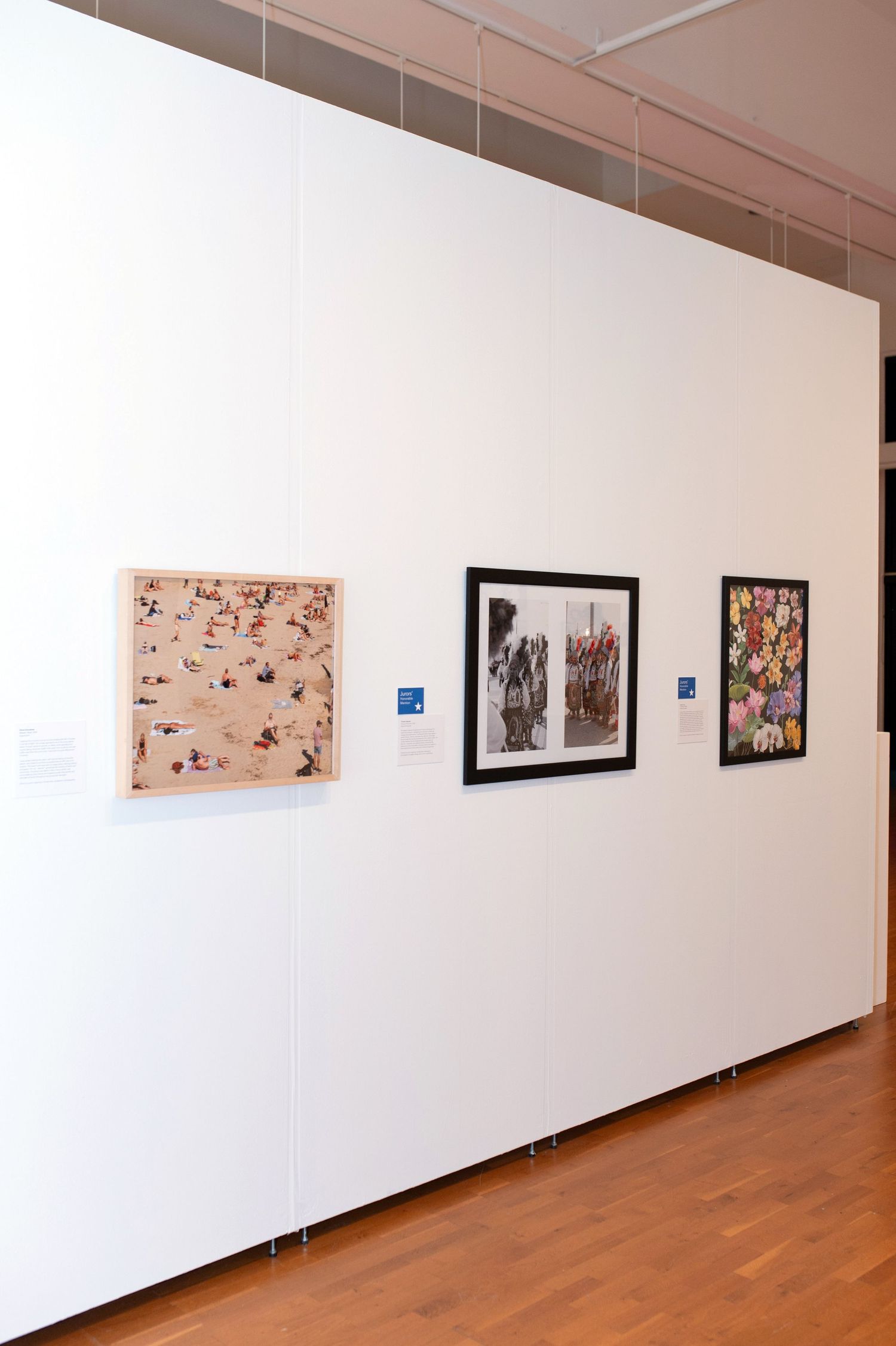 Three framed artworks displayed on white gallery wall with wooden flooring.