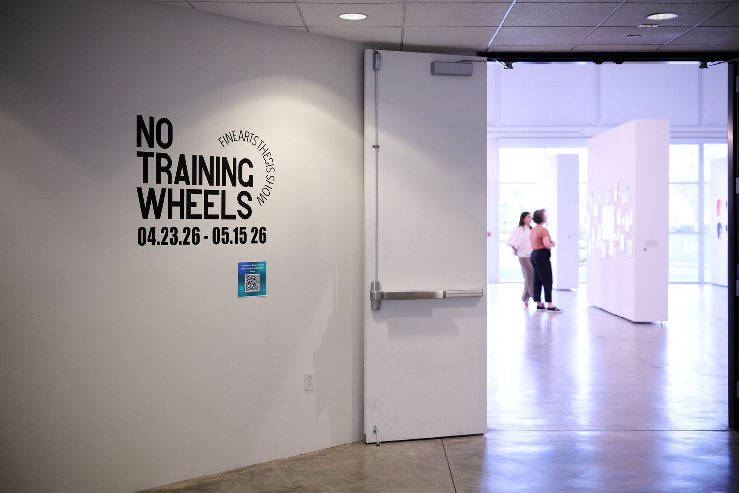 Gallery entrance with 'No Training Wheels' exhibition signage, dates 04.23.26-05.15.26, white walls, visitors inside.