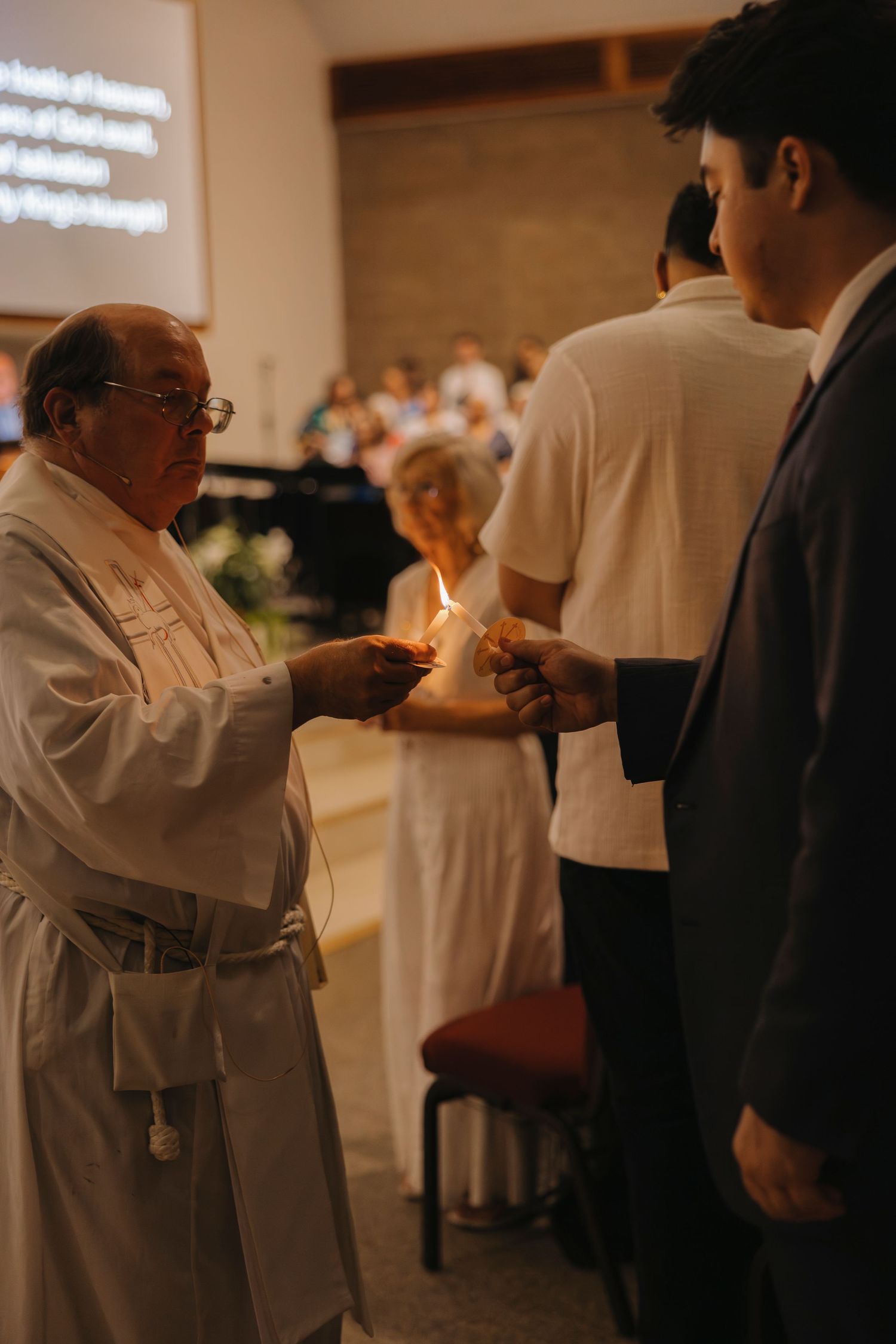 Priest in white robes sharing a candle flame with a young man during a church ceremony.
